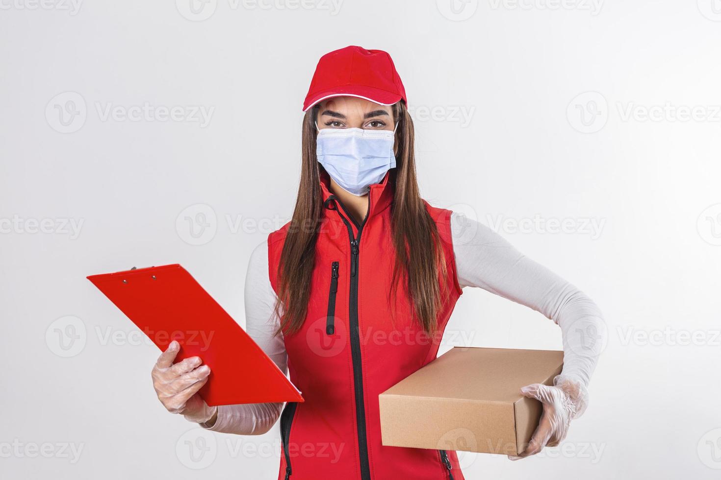 repartidor entregando paquetes con portapapeles y paquete sonriendo feliz en uniforme rojo. Hermosa mujer joven con máscara médica y guantes mensajero profesional aislado sobre fondo blanco. foto