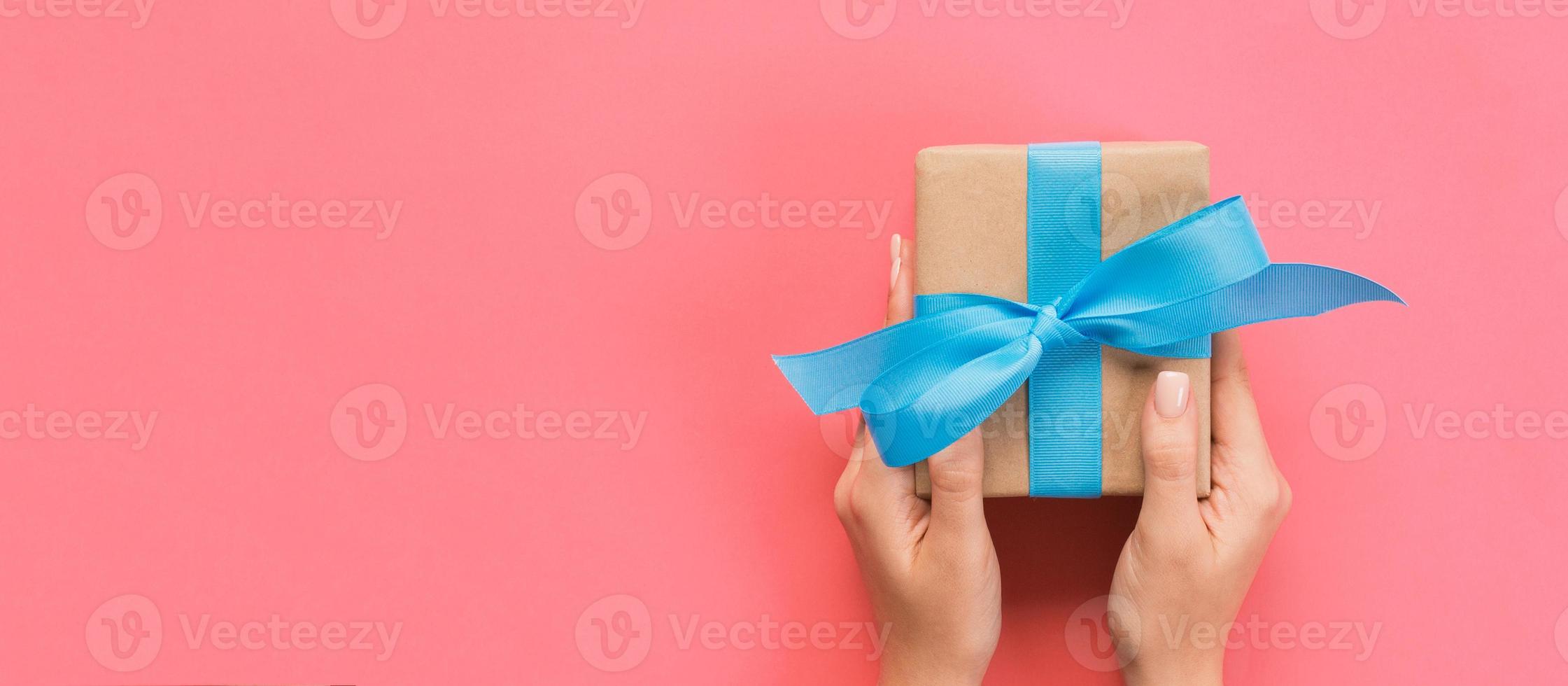 Woman hands give wrapped valentine or other holiday handmade present in paper with blue ribbon. Present box, decoration of gift on pink table, Top view banner with copy space for you design photo