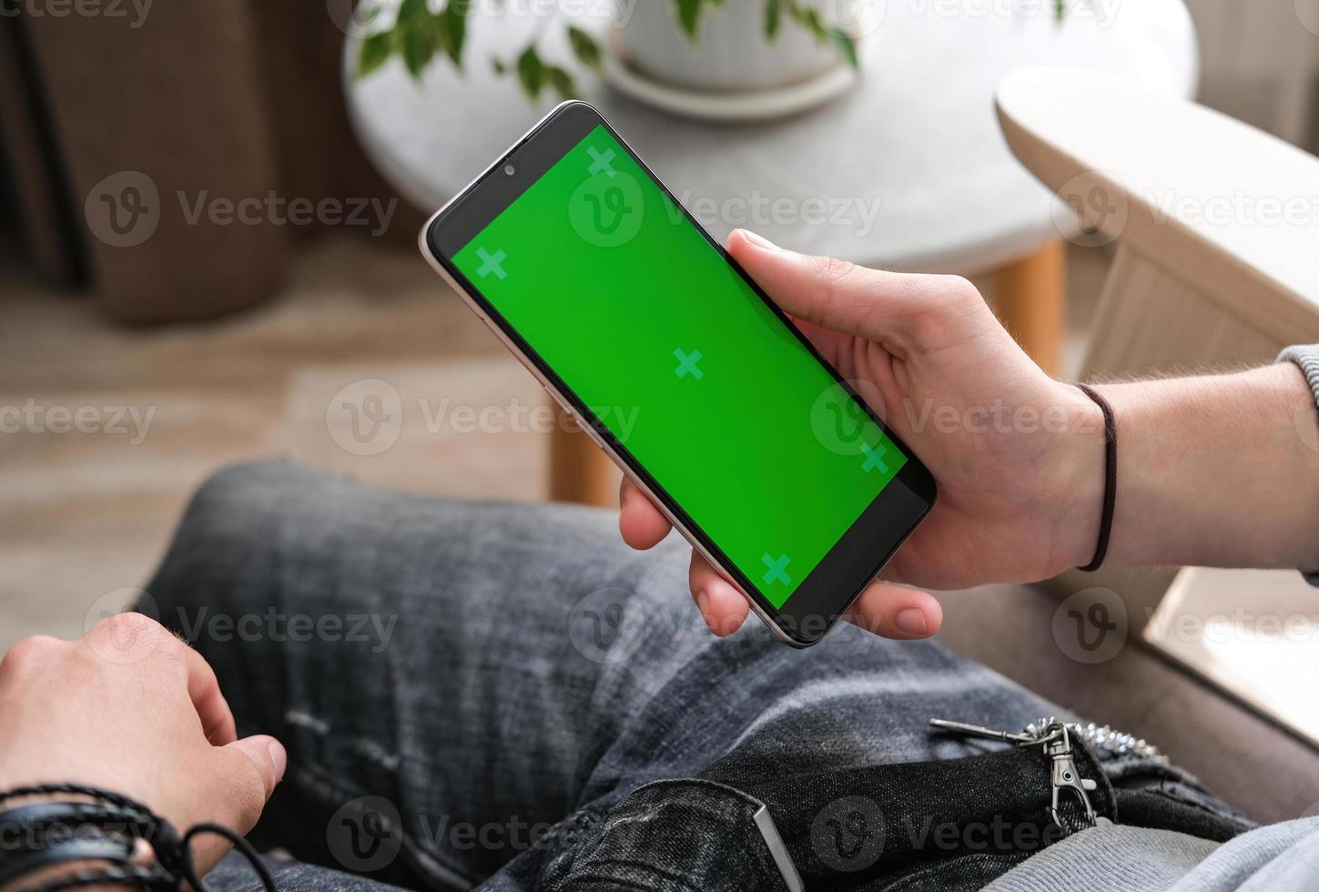 A young guy is looking at a phone screen with a chrome key indoors. Person using smartphone with green screen photo