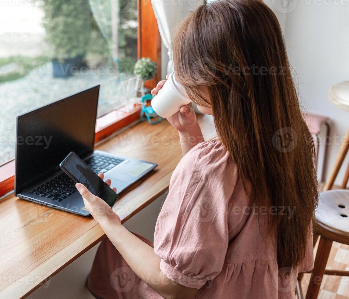 A happy  female at a cafe using a smartphone and laptop drinking coffee in a paper cup in the cafe. young white woman with long hair sitting in a coffee shop busy working on her smartphone. photo