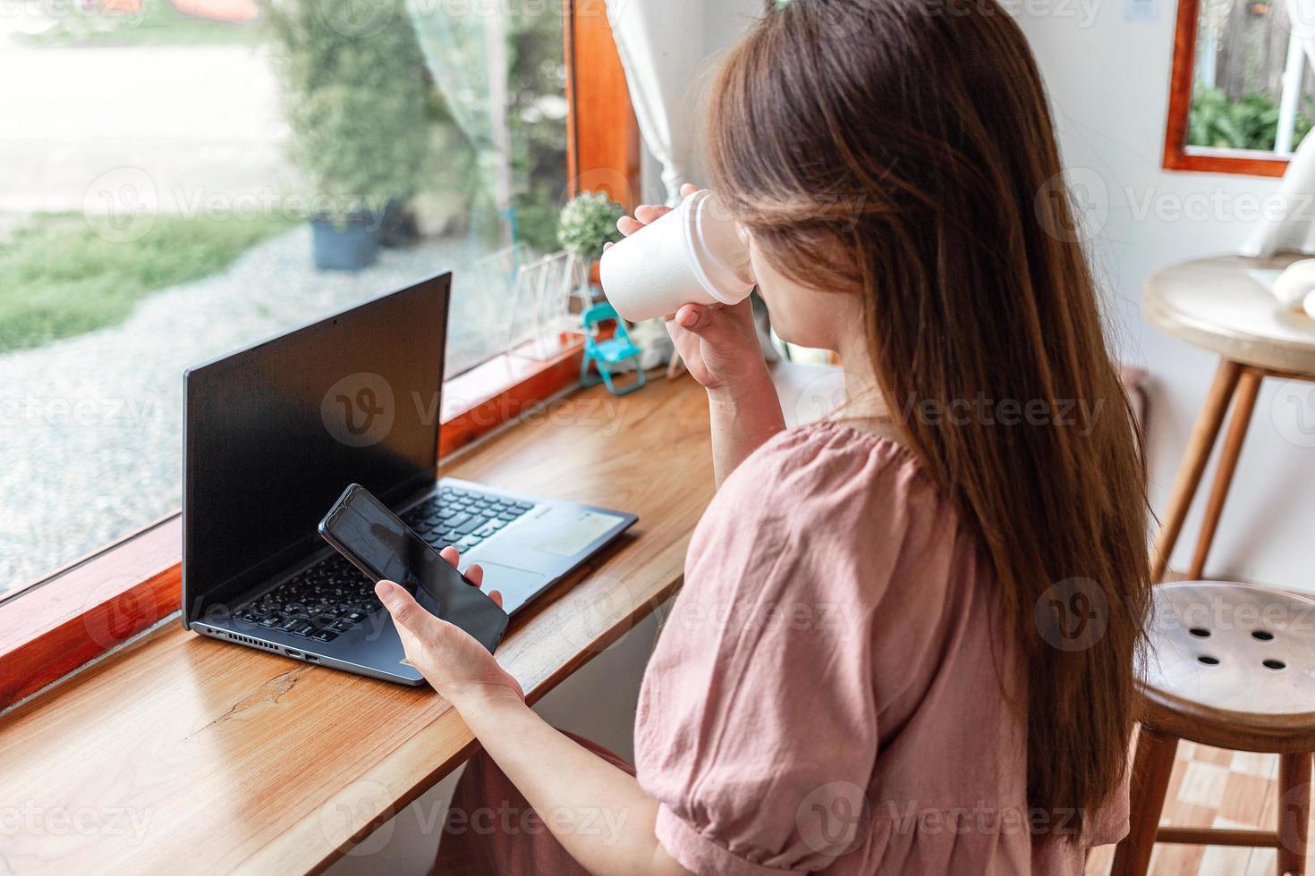 A happy female at a cafe using a smartphone and laptop drinking coffee in a paper cup in the cafe. young white woman with long hair sitting in a coffee shop busy working on her smartphone. photo