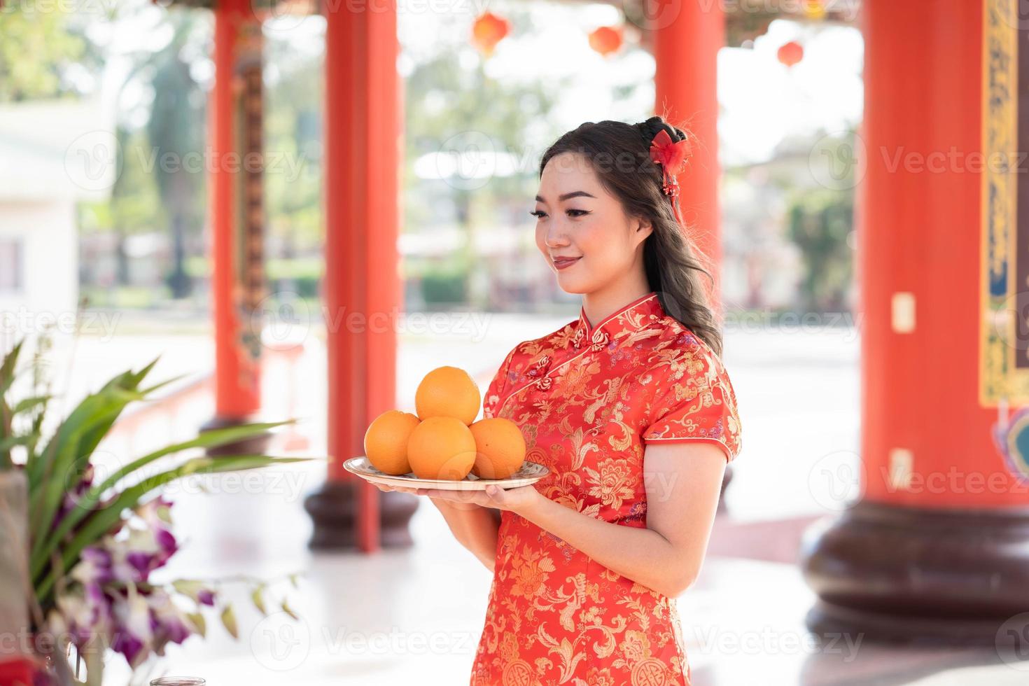 hermosa joven sonriendo alegremente usando el tradicional vestido cheongsam qipao sosteniendo naranjas frescas orando por la mejor bendición y buena suerte en el templo budista chino. concepto de año nuevo lunar foto