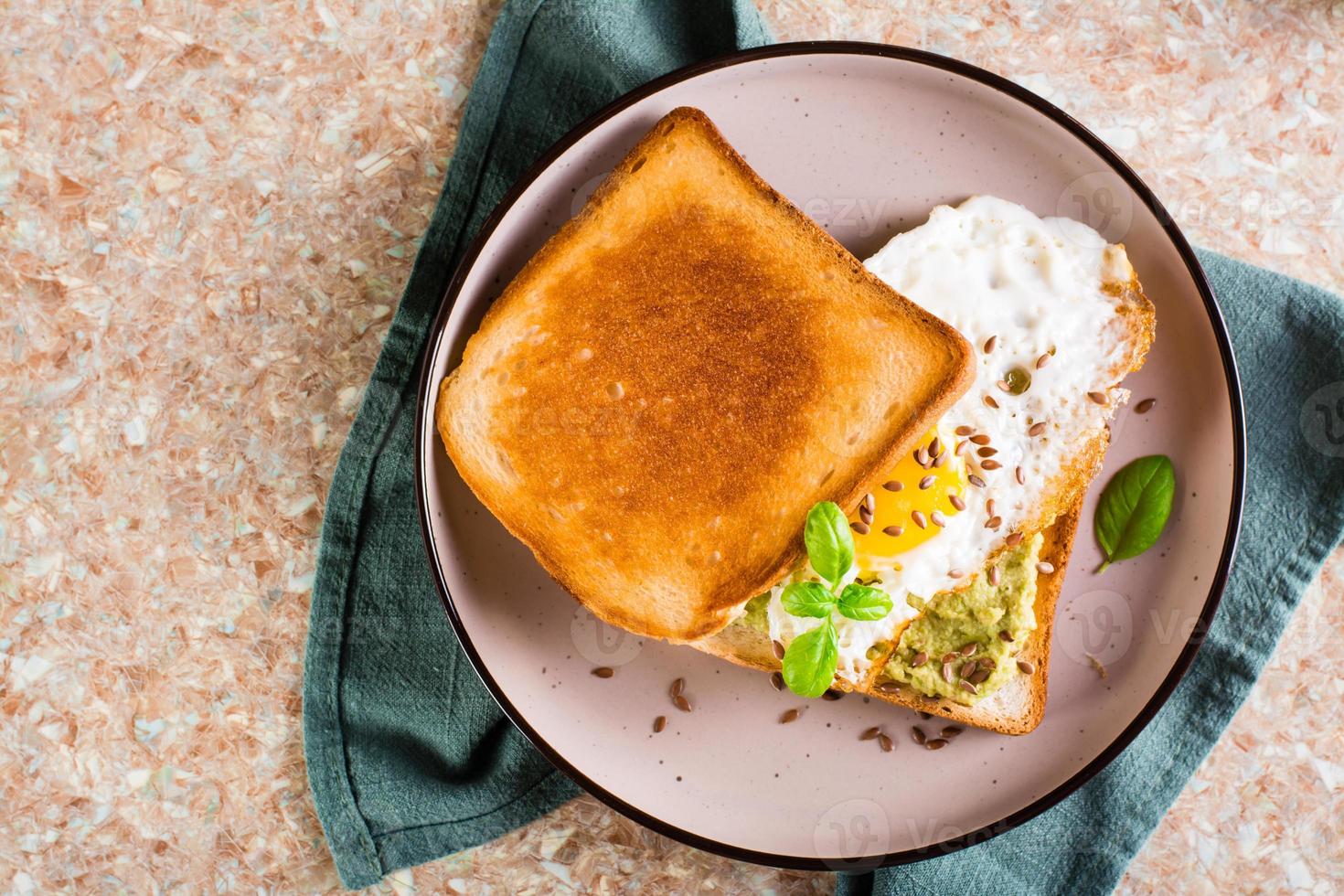 Sandwich with avocado, fried egg and flax seeds on toast on a plate on the table. Top view. Closeup photo