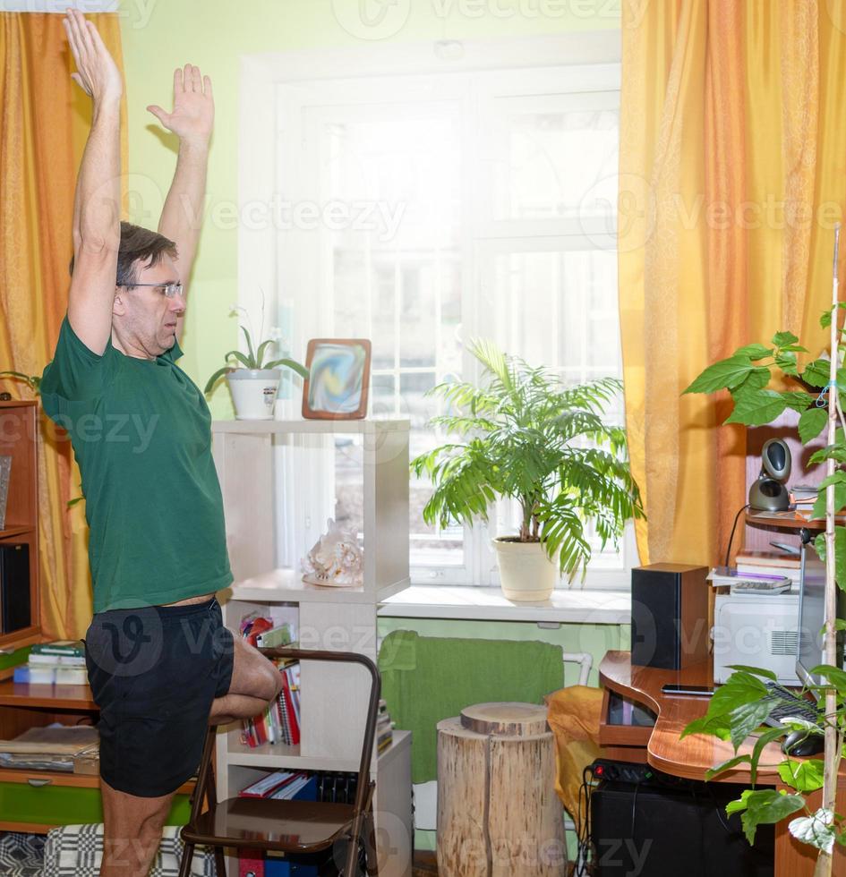 Mature man doing yoga asana pose tree before computer monitor in sunny room at home. Remote sport during pandemic. photo
