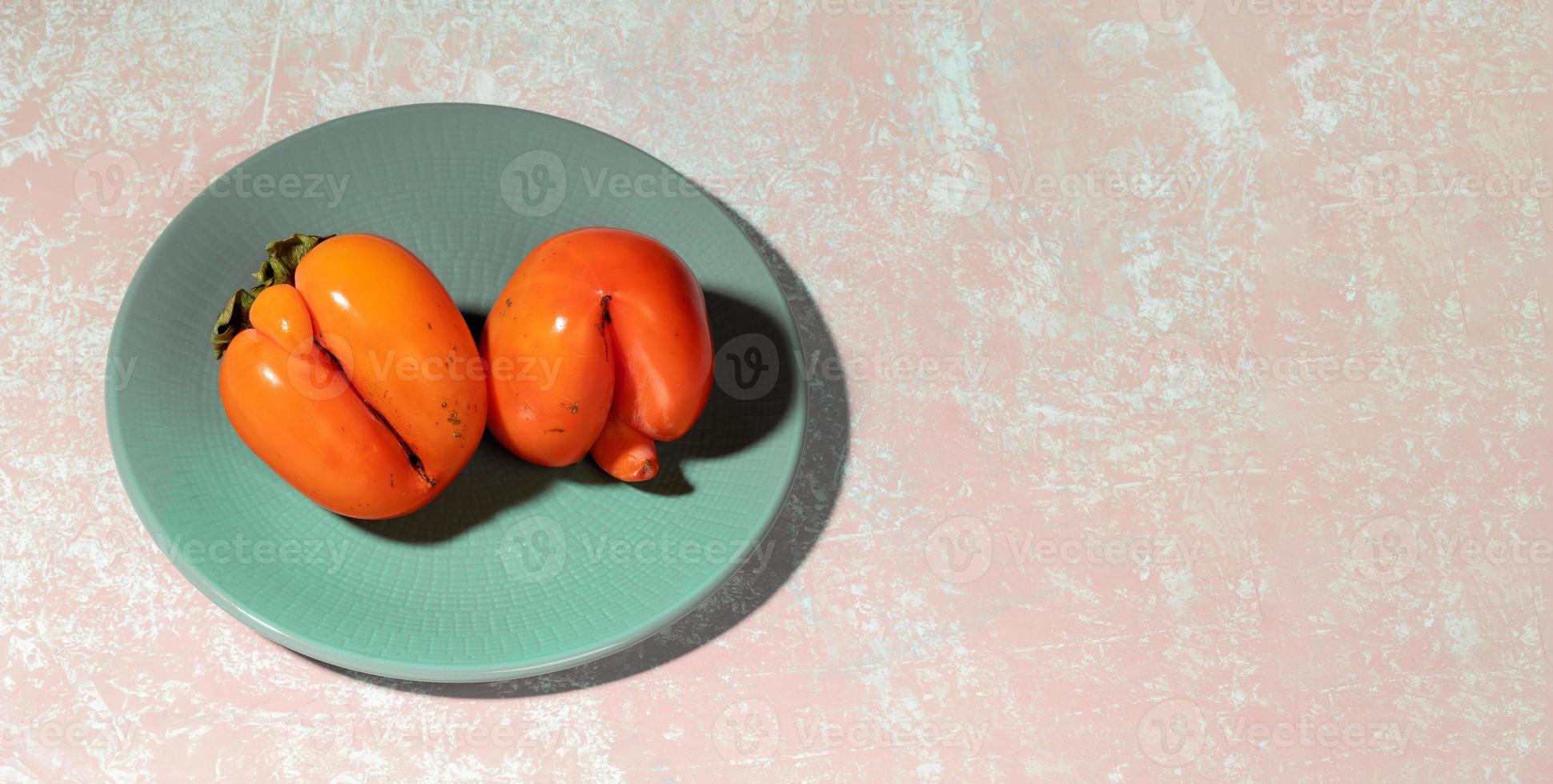 Banner with two ugly orange persimmons lying on turquoise plate on pink textured backdrop with copy space. photo