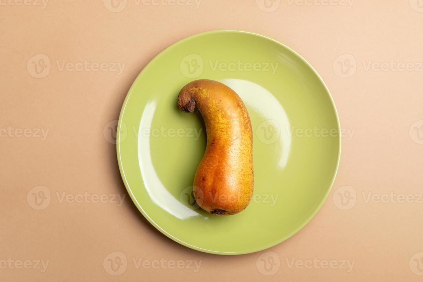 View from above to one ugly crooked ripe pear lying on round green plate on beige background. photo