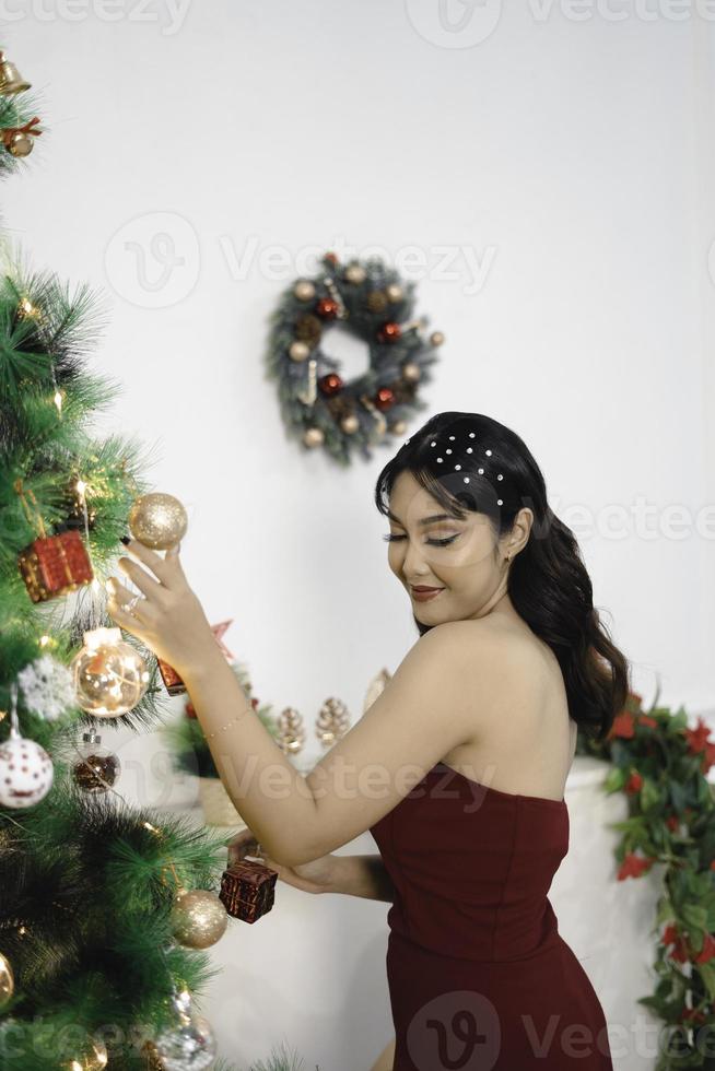 Portrait of pretty young girl decorating Christmas tree, smiling wearing red gown in decorated Christmas living room indoors photo