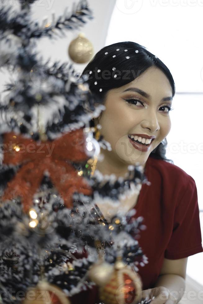 Portrait of pretty young girl decorating Christmas tree, smiling wearing red gown in decorated Christmas living room indoors photo