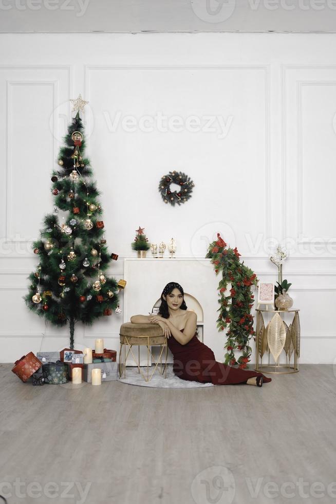 retrato de una joven bonita y acogedora sentada en el suelo, sonriendo, vestida con un vestido rojo en una sala de estar navideña decorada en el interior foto