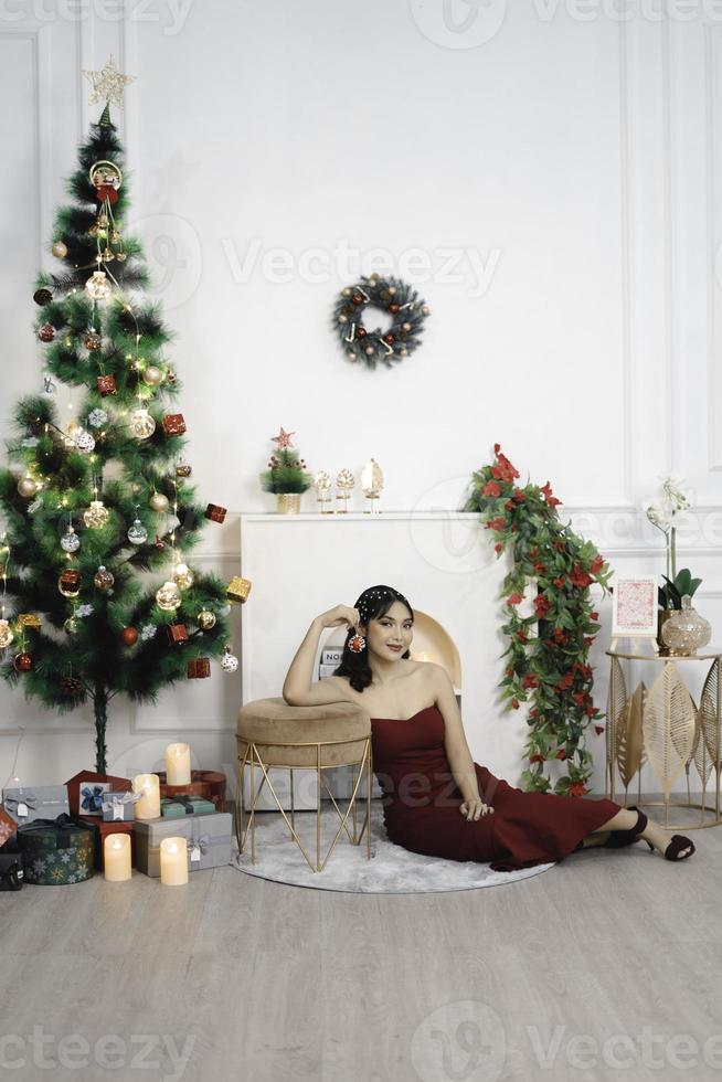 retrato de una joven bonita y acogedora sentada en el suelo, sonriendo, vestida con un vestido rojo en una sala de estar navideña decorada en el interior foto