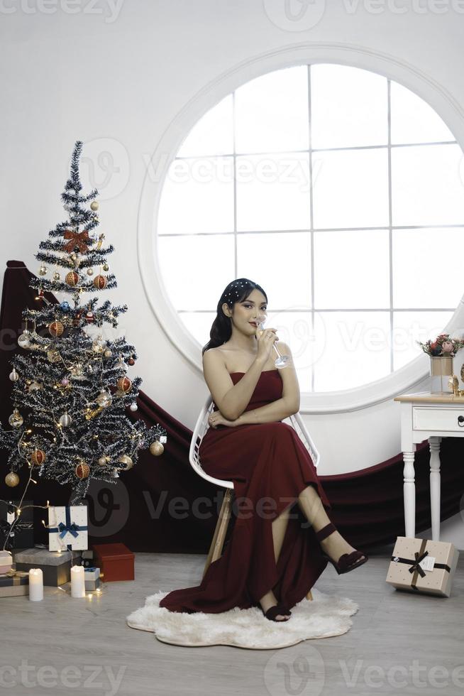 Portrait of pretty young girl cozy sit down and drinking wine, wear red gown in decorated Christmas living room indoors photo