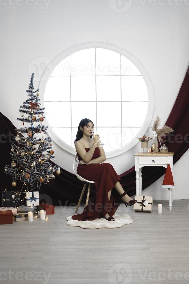 Portrait of pretty young girl cozy sit down and drinking wine, wear red gown in decorated Christmas living room indoors photo