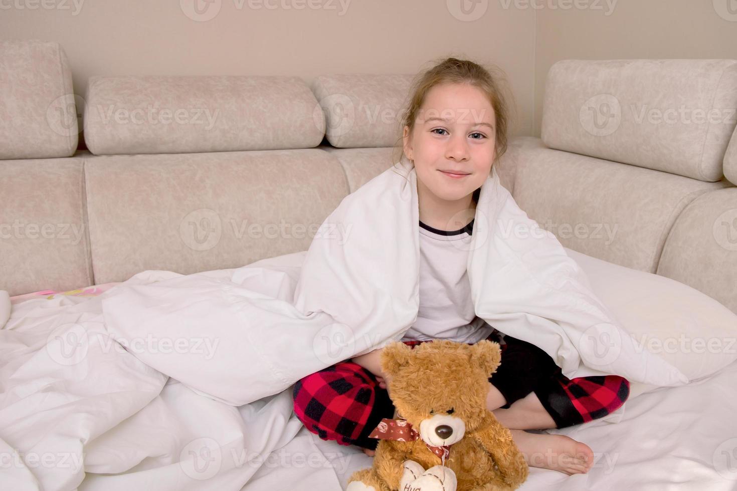 girl sits in bed in pajamas covered with a blanket with a bear and smiles photo