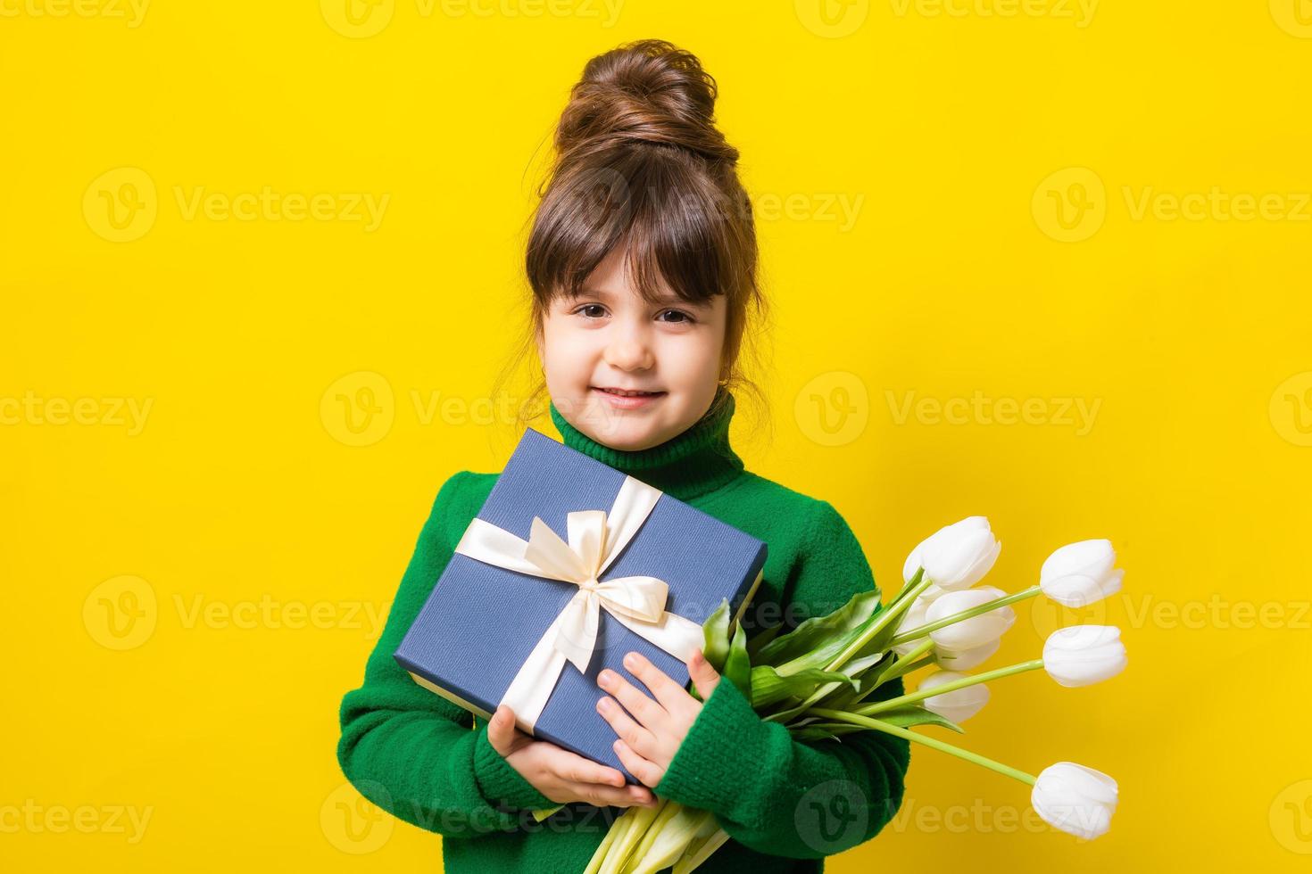 a happy little brunette girl is holding a gift box and a bouquet of tulips on a yellow background in the studio. The concept of Mother's Day, March 8, Valentine's Day. High quality photo