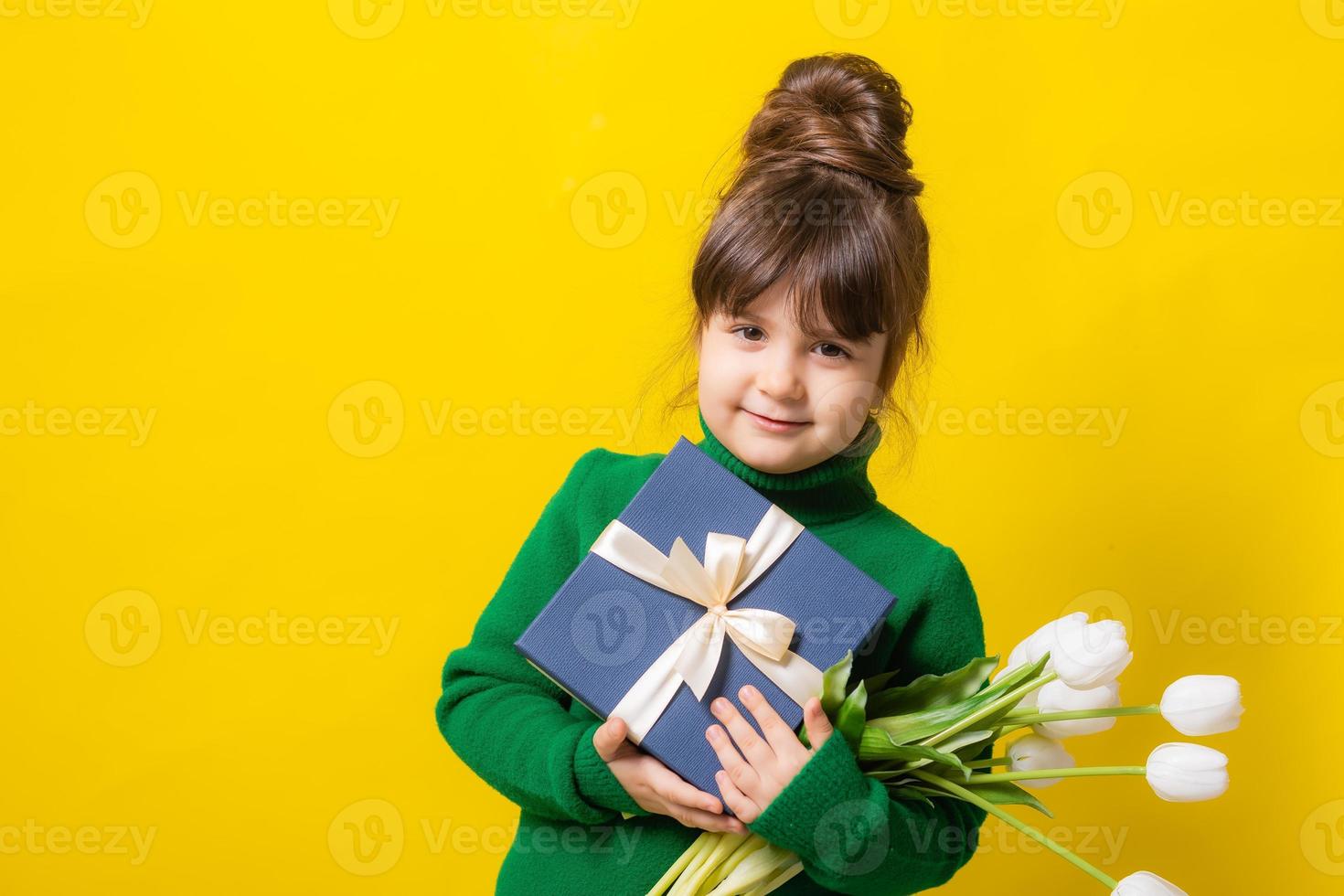 a happy little brunette girl is holding a gift box and a bouquet of tulips on a yellow background in the studio. The concept of Mother's Day, March 8, Valentine's Day. High quality photo