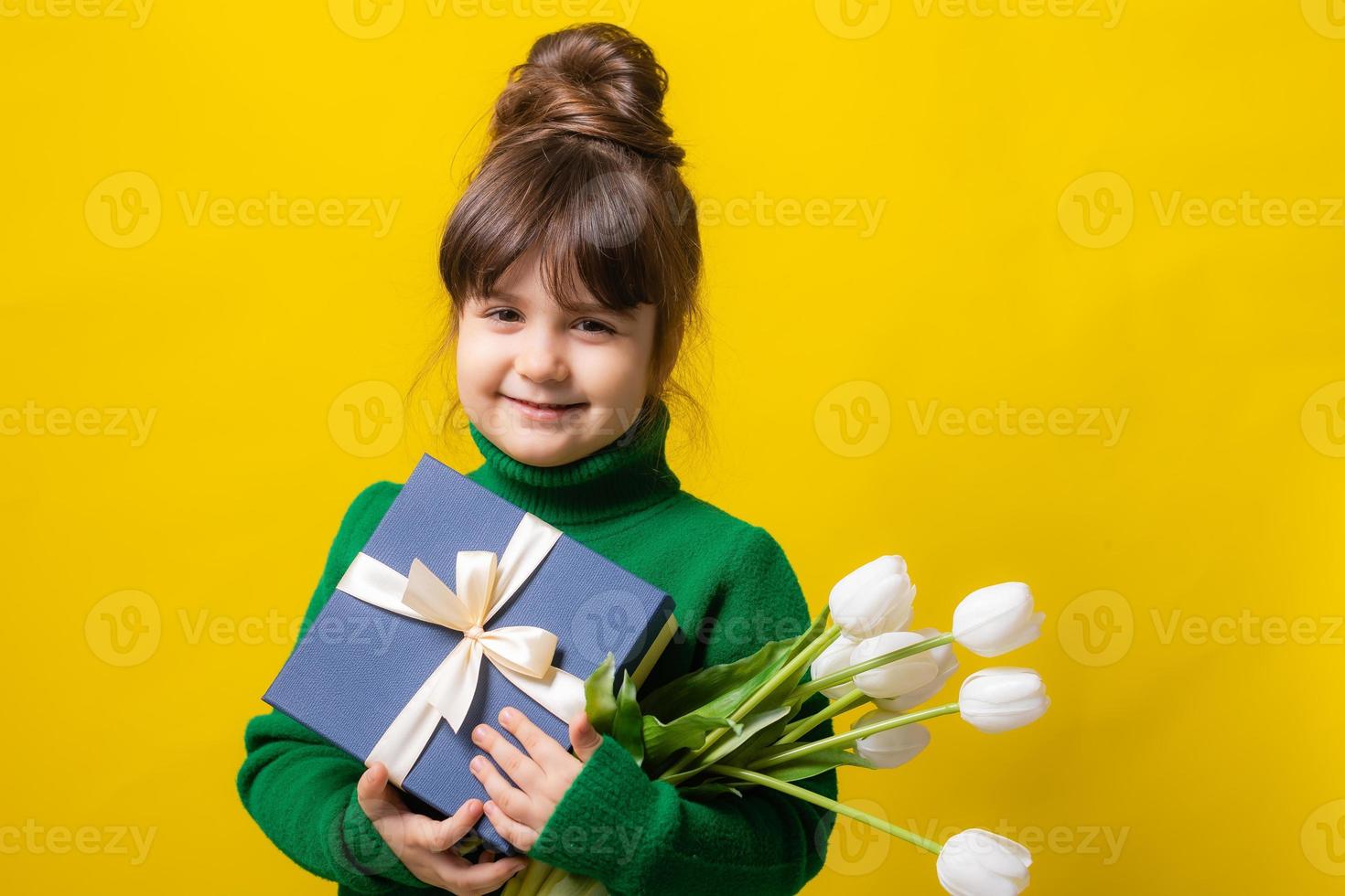 a happy little brunette girl is holding a gift box and a bouquet of tulips on a yellow background in the studio. The concept of Mother's Day, March 8, Valentine's Day. High quality photo