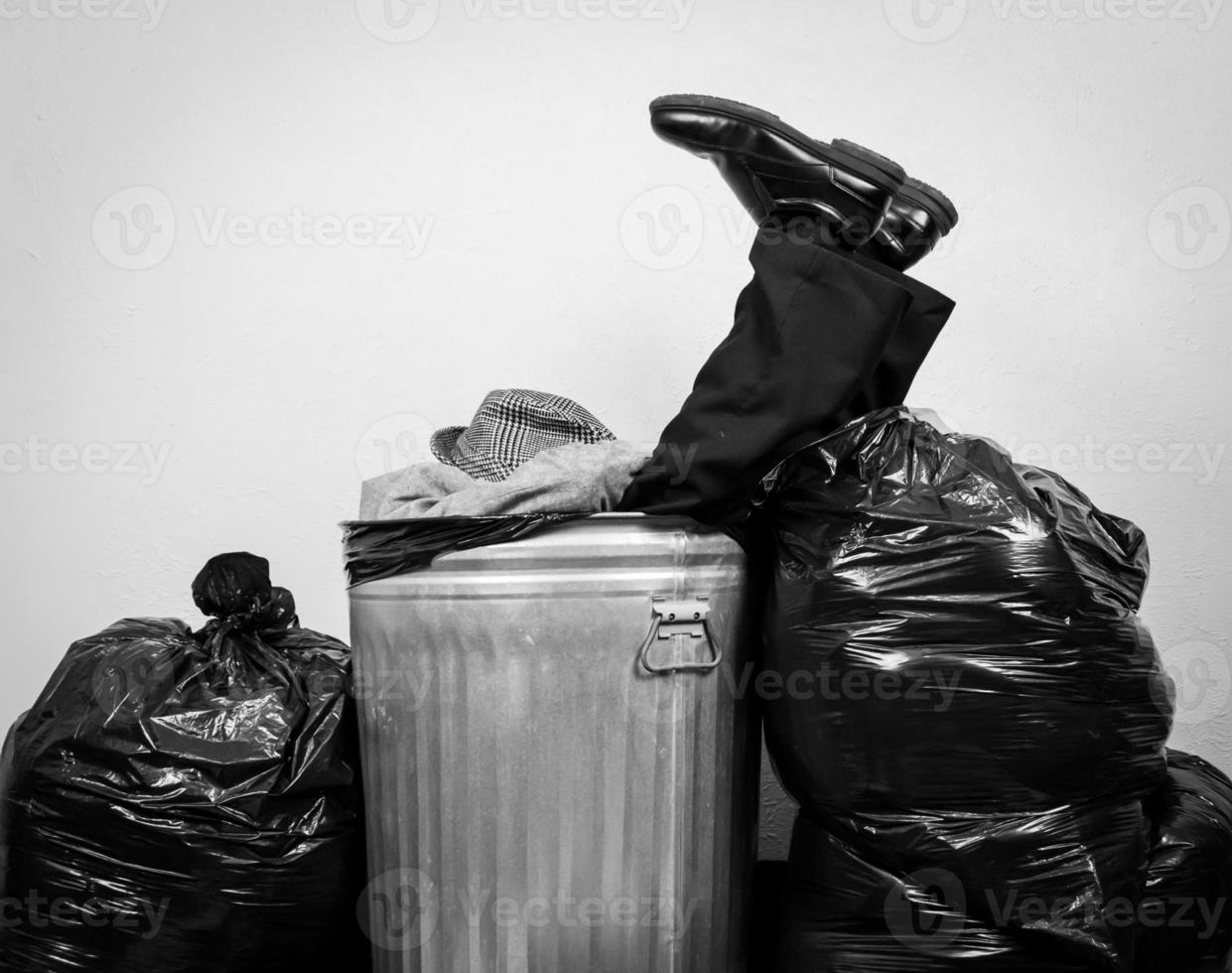 retrato en blanco y negro de un hombre de negocios con traje sentado en un bote de basura de metal rodeado de bolsas de basura. concepto de hombre golpeado por el capitalismo y la vida. foto