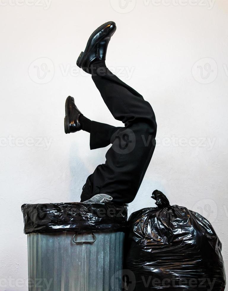 Businessman in Suit Stuck Upside Down in Metal Trash Can Next to Garbage Bag Pile. Concept of Over a Barrel. Thrown Away by Capitalism and Greed. photo