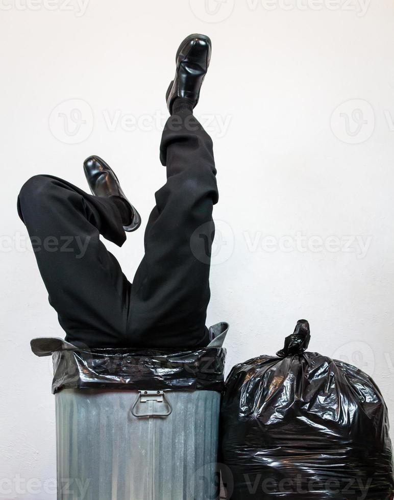 hombre de negocios con traje atrapado boca abajo en un bote de basura de metal junto a la pila de bolsas de basura. concepto de más de un barril. desechados por el capitalismo y la codicia. foto