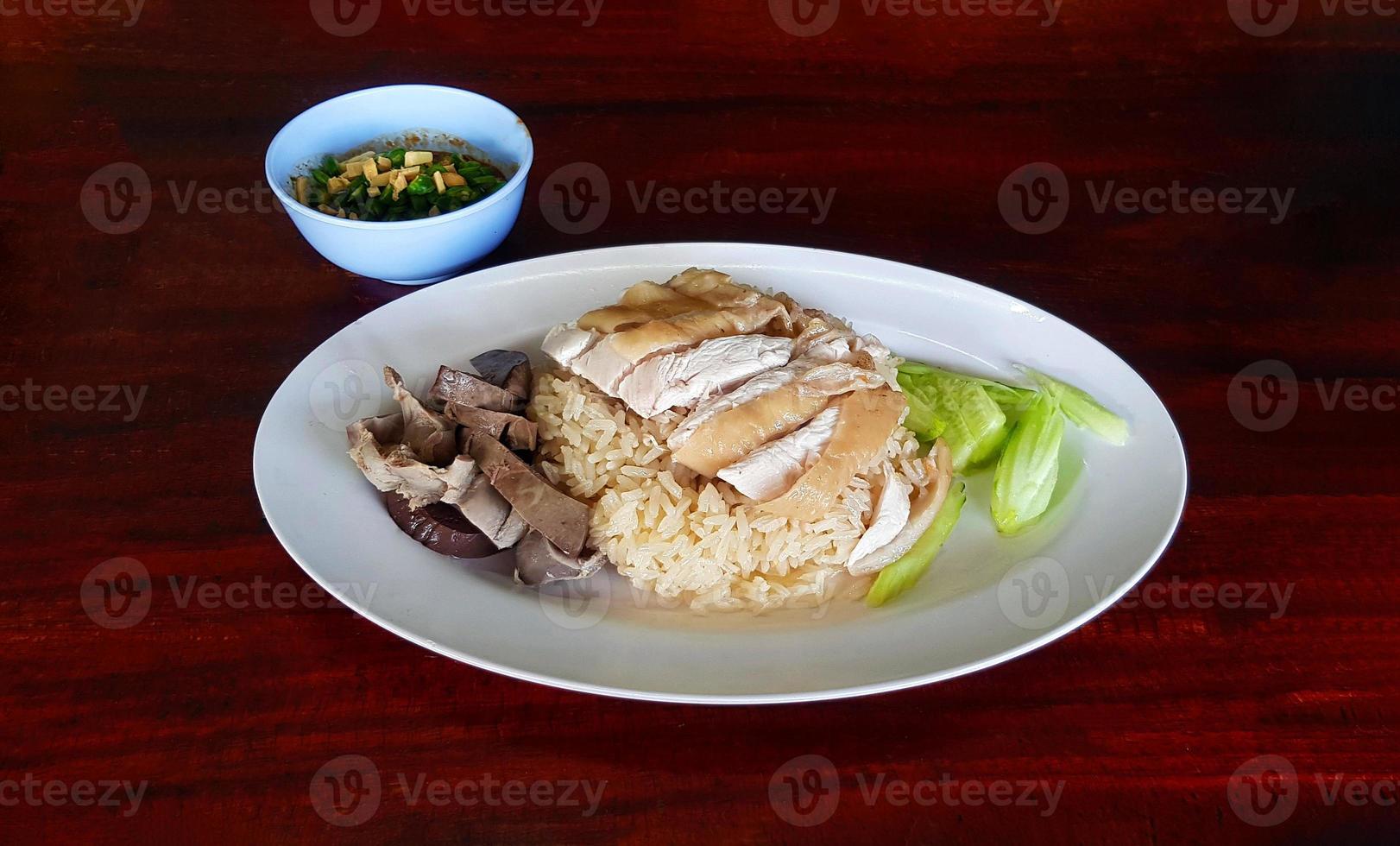 Boiled chicken, offal with cucumber sliced on white dish and small bowl spicy or sweet sauce on dark brown wooden background. Steamed rice topped with chicken and fresh vegetable on table. Asian food. photo