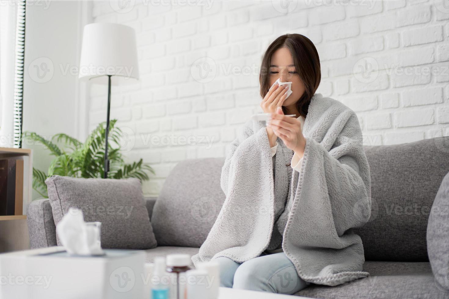 Sick young asian woman sit under blanket on sofa. Female blow nose, coughing, sneezing, check temp in tissue at home, suffering from flu. photo