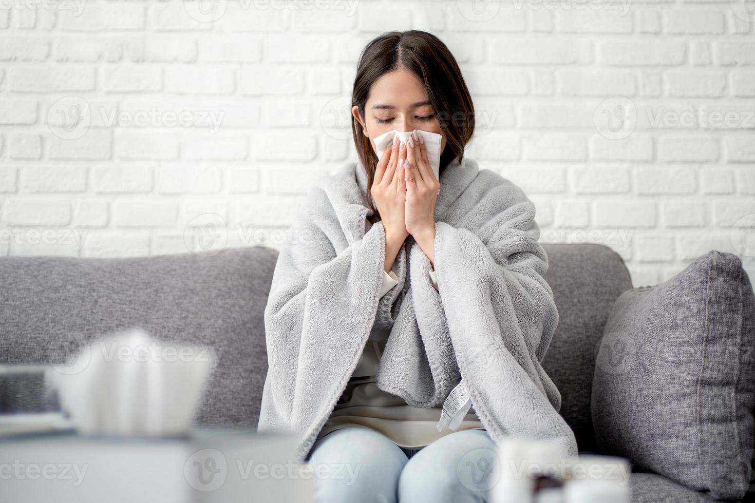 mujer asiática enferma cubierta de manta estornudando sentada en el sofá estaba enferma y tenía fiebre. concepto de gripe foto