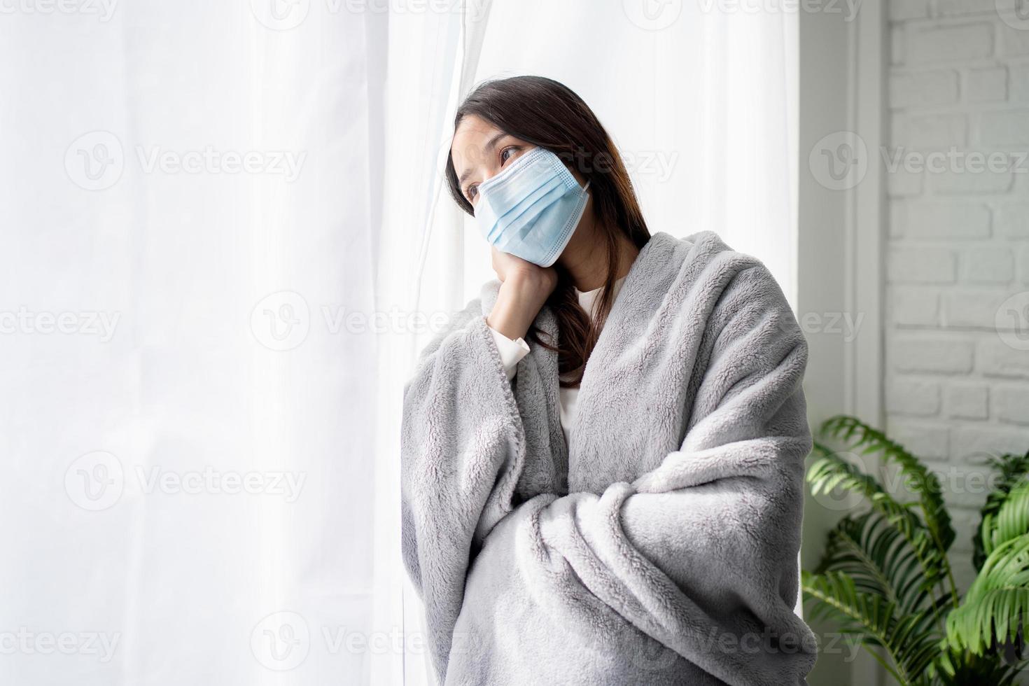 triste niña asiática enferma en cuarentena, hermosa mujer asiática parada cerca de la ventana en casa y mirando afuera, ella usa mascarilla médica de covid foto