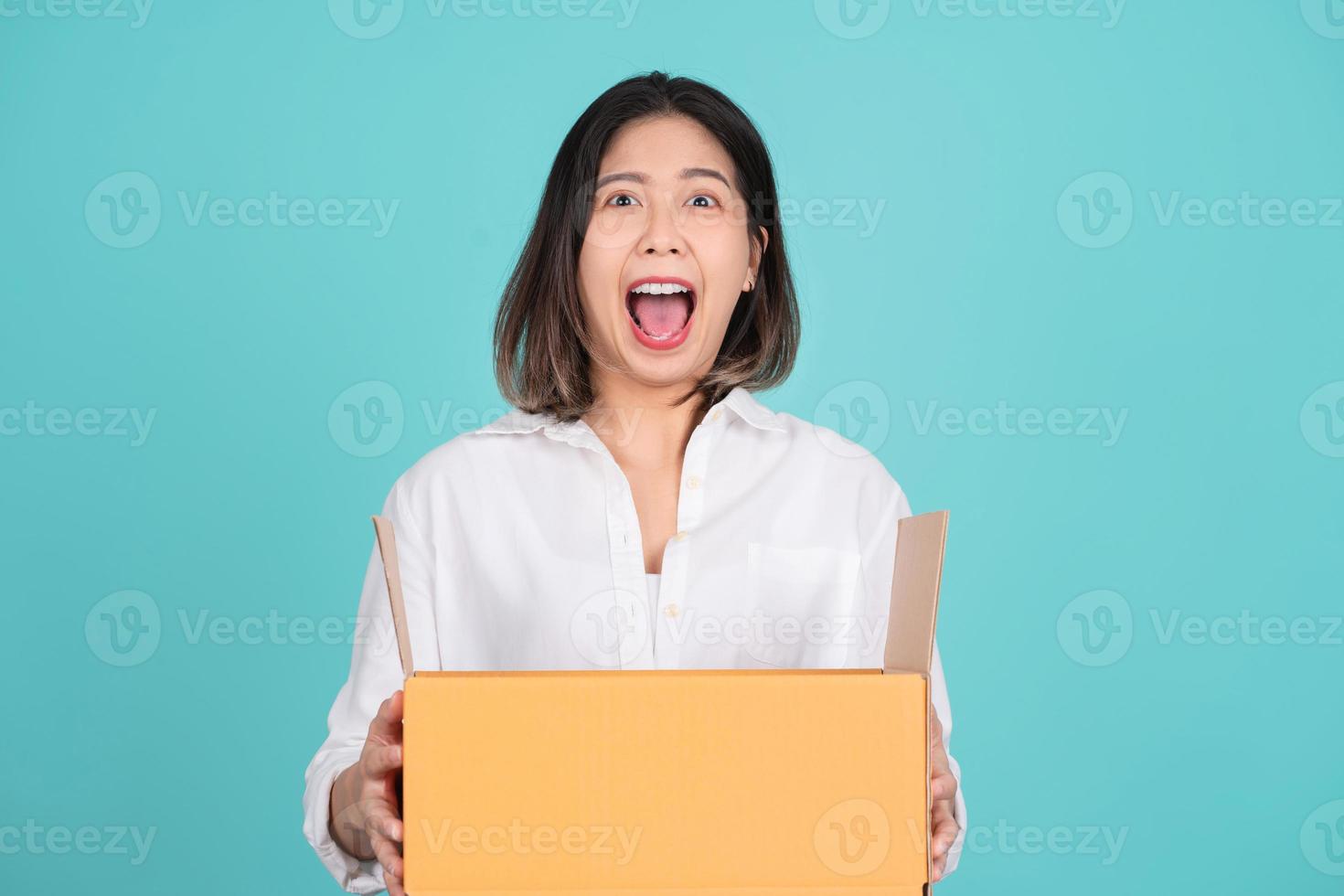 Shocked Asian woman is holding cardboard box on turquoise background. photo