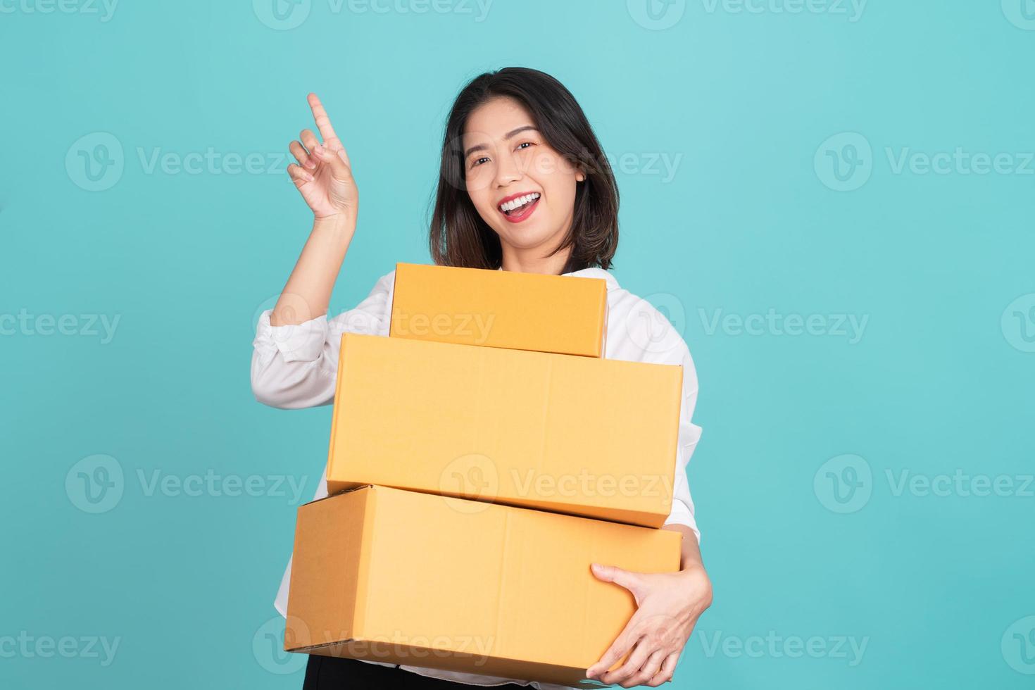 Happy Asian woman holding package parcel box and pointing finger to empty copy space isolated on light green background, photo