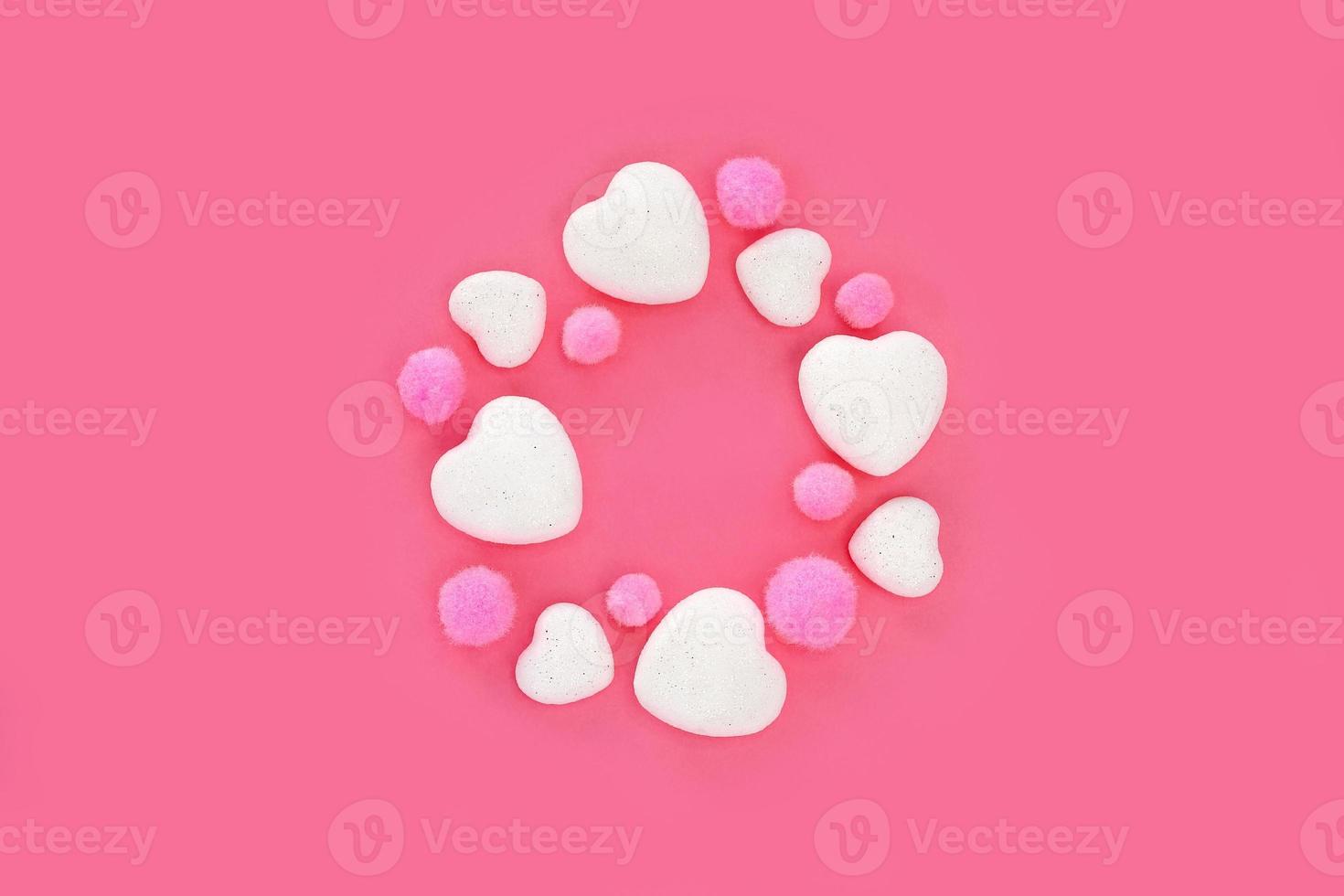 Valentine's Day decorations on pink background. White glitter hearts and pink fluffy pompoms arranged in a circle, centered on bright pink paper. Greeting card with copy space. Top view, flat lay. photo