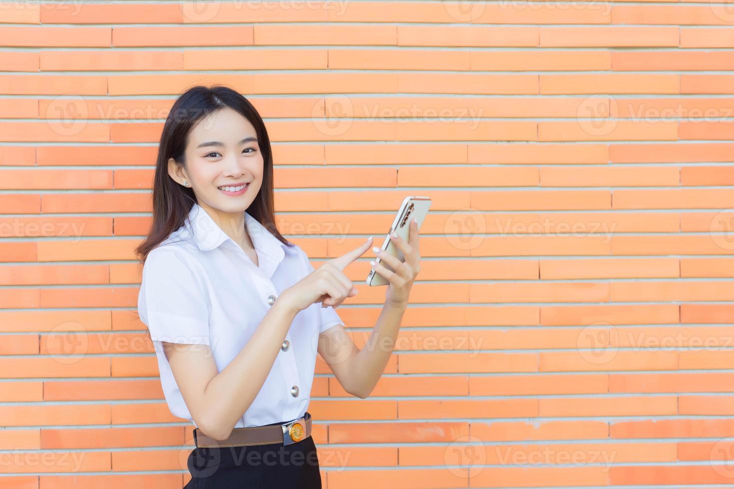 Portrait of cute Asian Thai girl student in uniform is standing smiling happily and confidently successful while holds smartphone with brick walls as background in university. photo