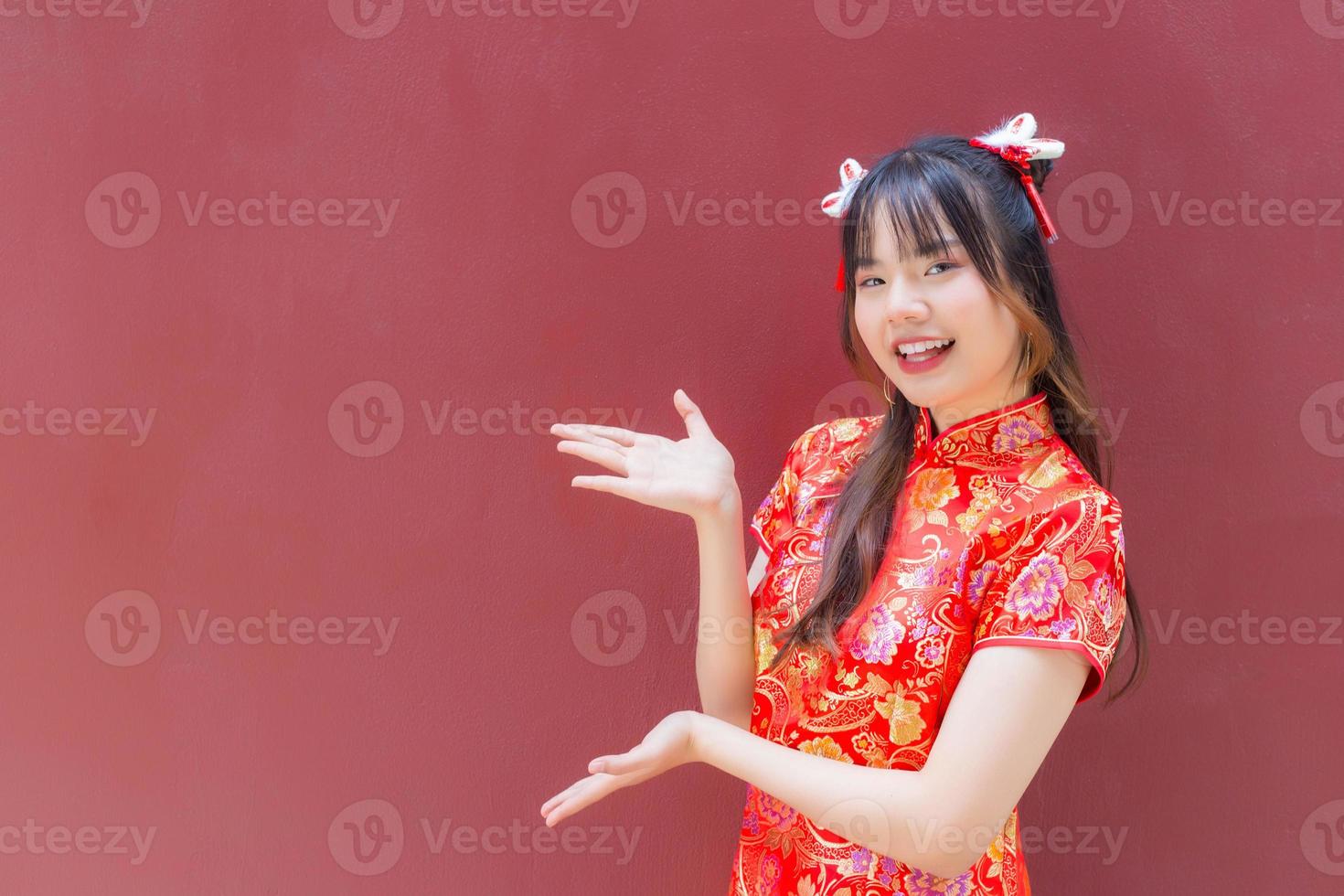 linda chica asiática con el pelo largo que usa un vestido rojo cheongsam en el tema del año nuevo chino mientras muestra su mano para presentar algo sobre un fondo rojo. foto