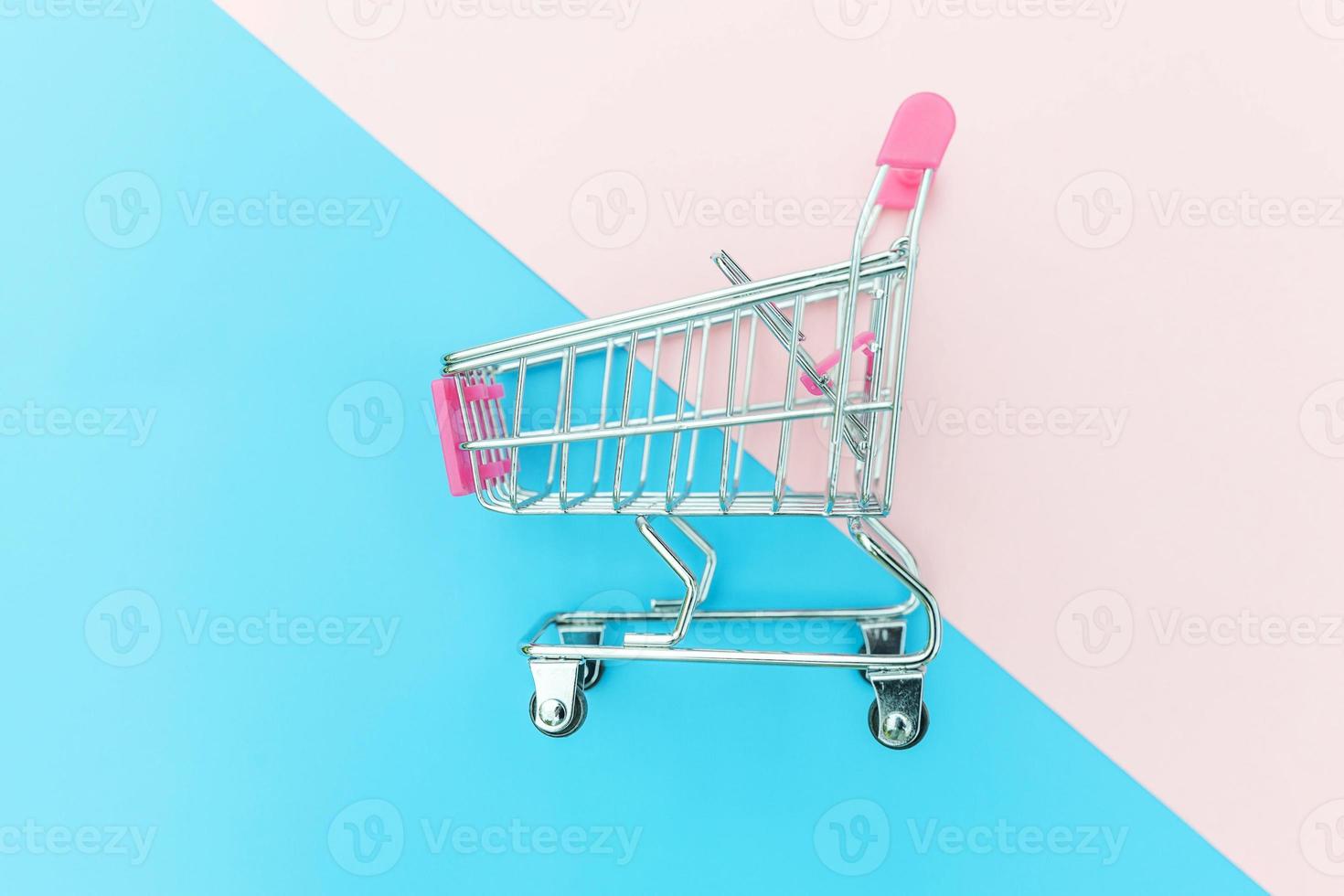 Small supermarket grocery push cart for shopping toy with wheels isolated on blue and pink pastel colorful trendy geometric background Copy space. Sale buy mall market shop consumer concept. photo