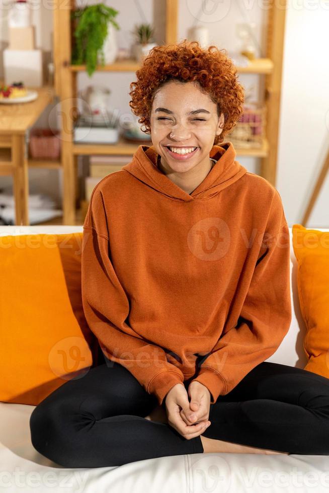 Beautiful african american girl with afro hairstyle smiling sitting on sofa at home indoor. Young african woman with curly hair laughing. Freedom happiness carefree happy people concept. photo