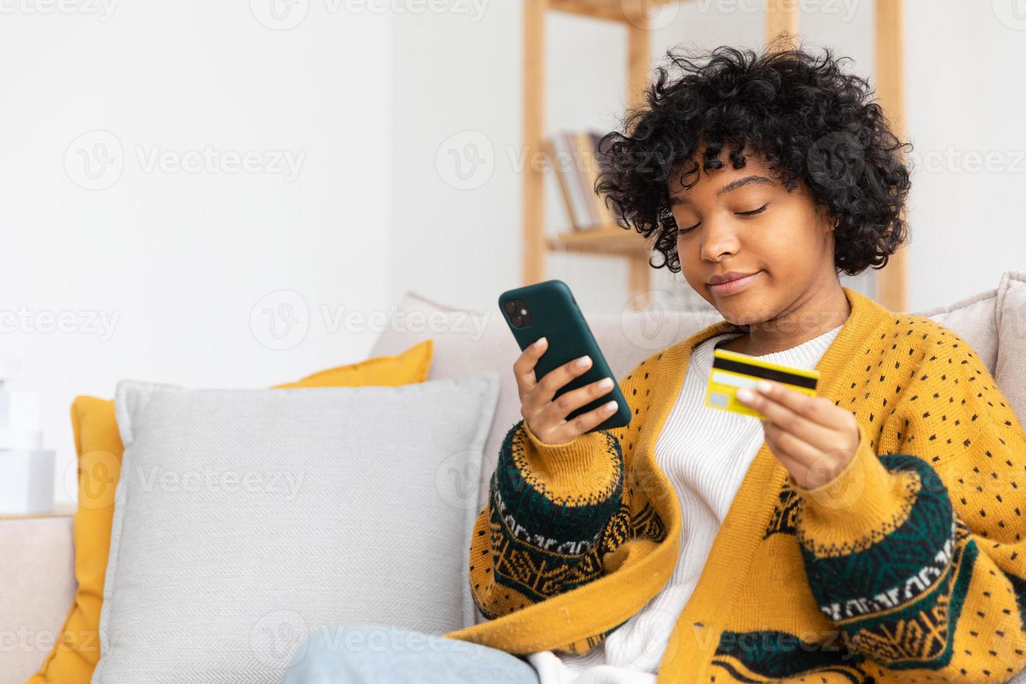 African american woman shopping online holding smartphone paying with gold credit card. Girl sitting at home buying on Internet enter credit card details. Online shopping ecommerce delivery service. photo