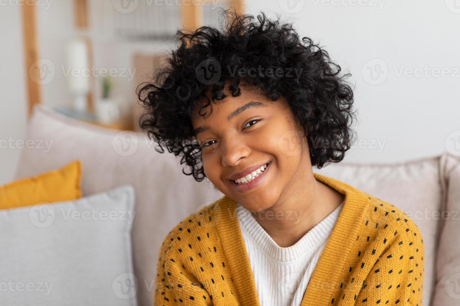 Beautiful african american girl with afro hairstyle smiling sitting on sofa at home indoor. Young african woman with curly hair laughing. Freedom happiness carefree happy people concept. photo