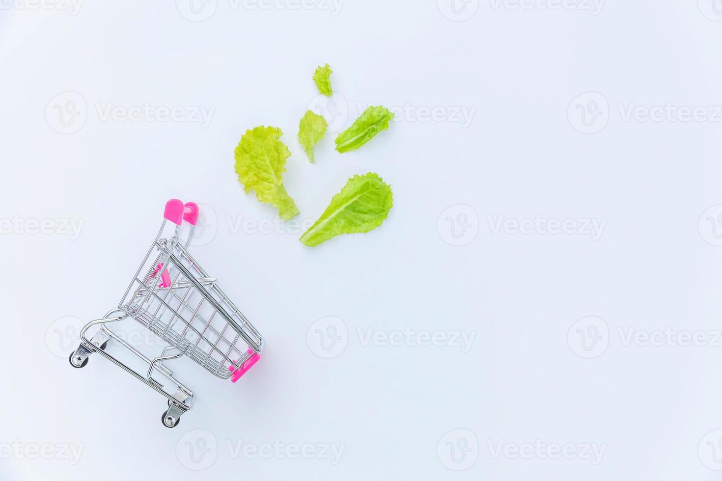 Ecology eco products health food vegan vegetarian concept . Small supermarket grocery push cart for shopping with green lettuce leaves isolated on white background. Flat lay top view copy space. photo