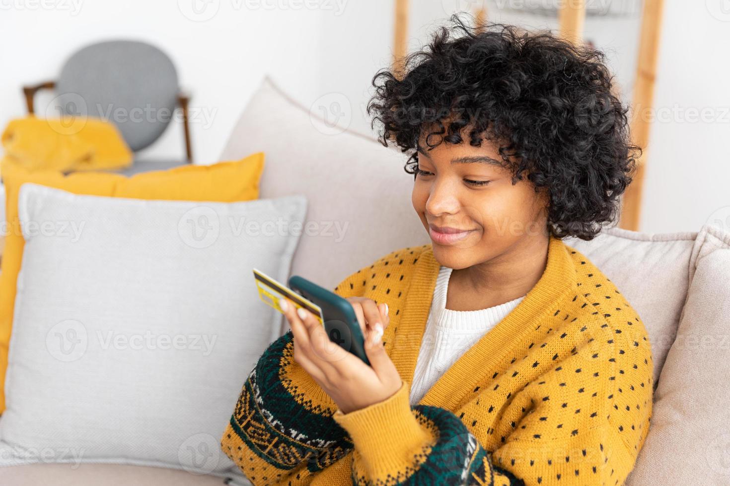 African american woman shopping online holding smartphone paying with gold credit card. Girl sitting at home buying on Internet enter credit card details. Online shopping ecommerce delivery service. photo