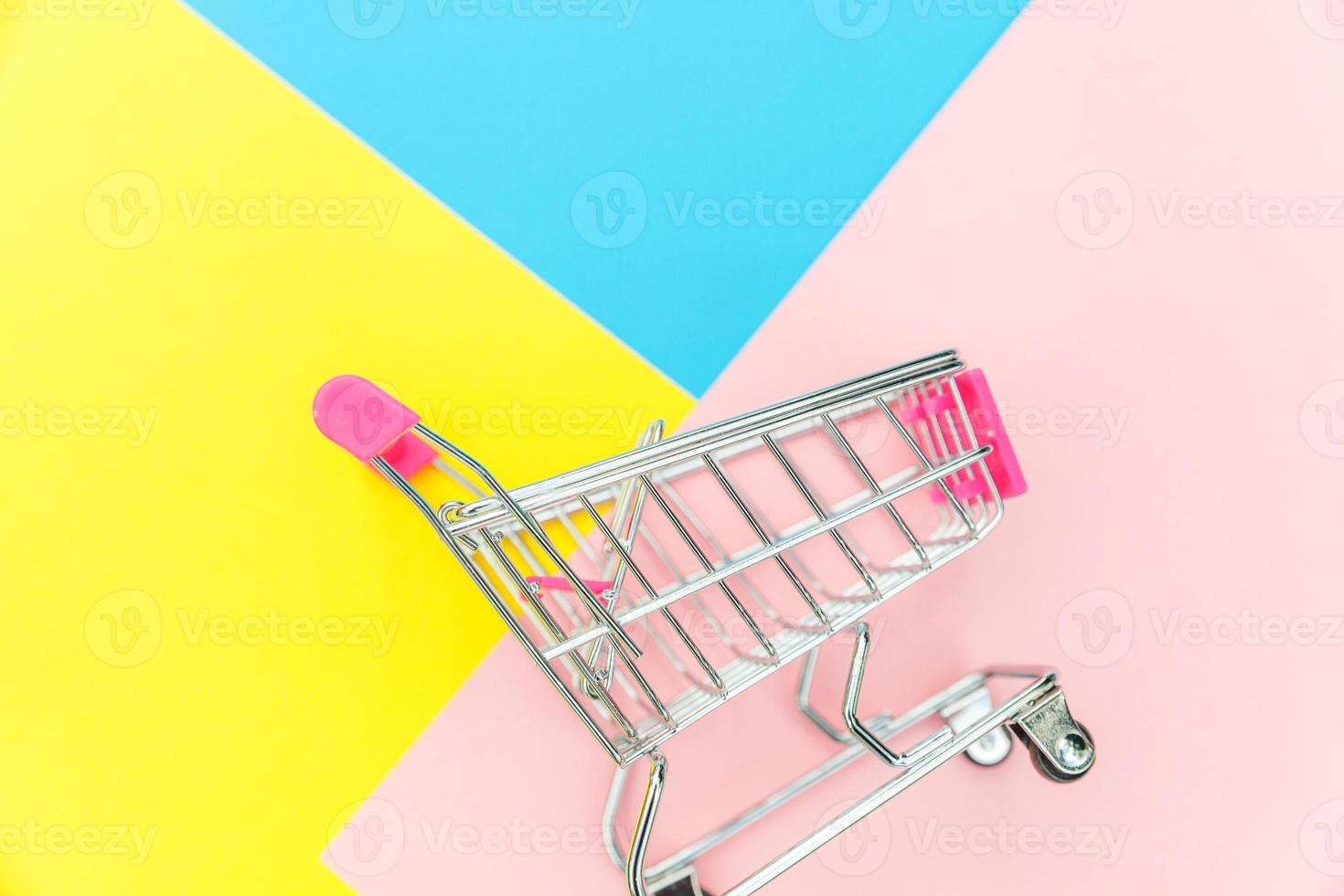 Small supermarket grocery push cart for shopping toy with wheels isolated on blue yellow pink pastel colorful trendy geometric background Copy space. Sale buy mall market shop consumer concept. photo