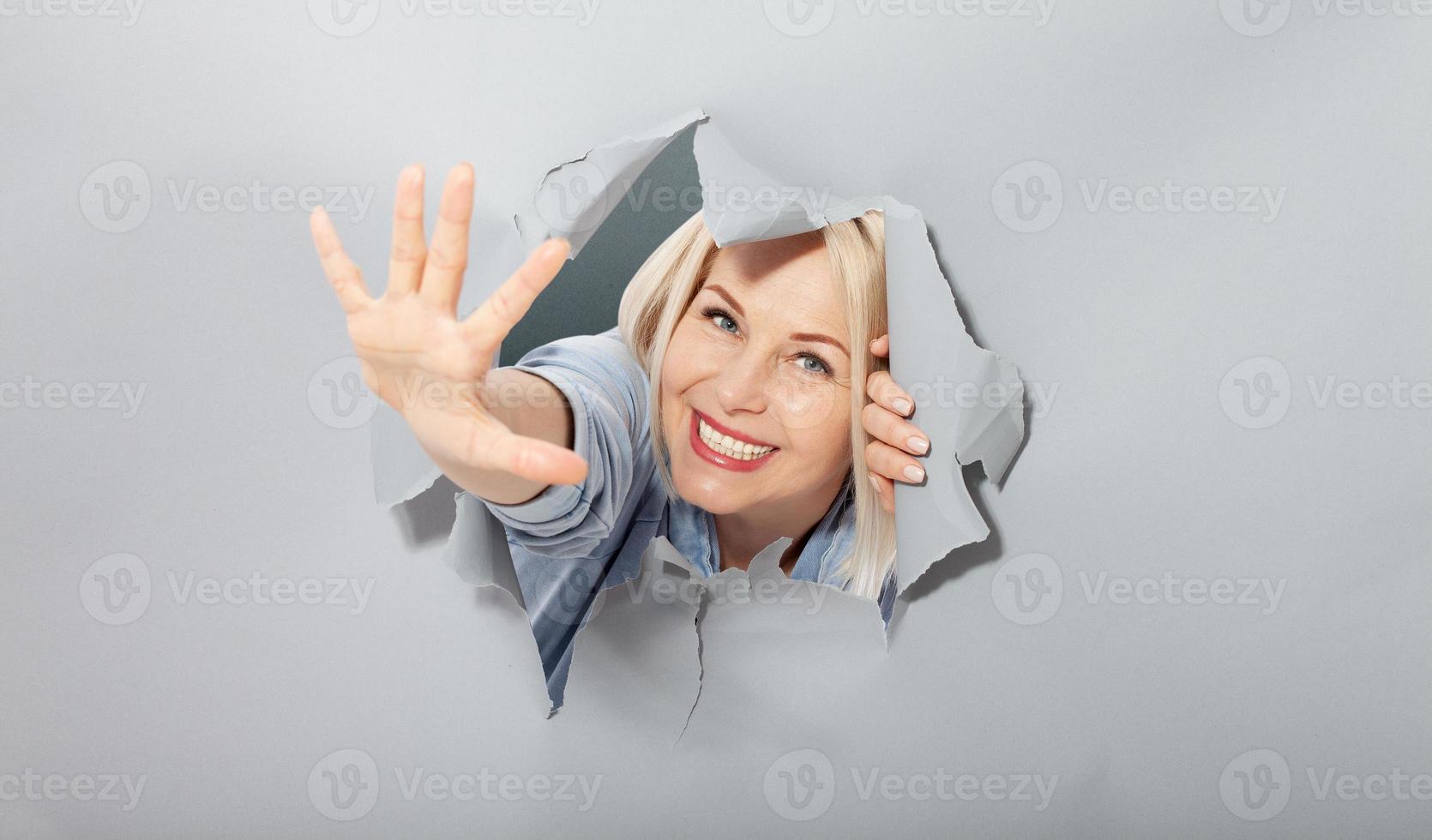 Attractive joyful middle aged woman waving a greeting gesture with her hand, shows that everything is ok everything is in order. Torn hole in gray paper. photo