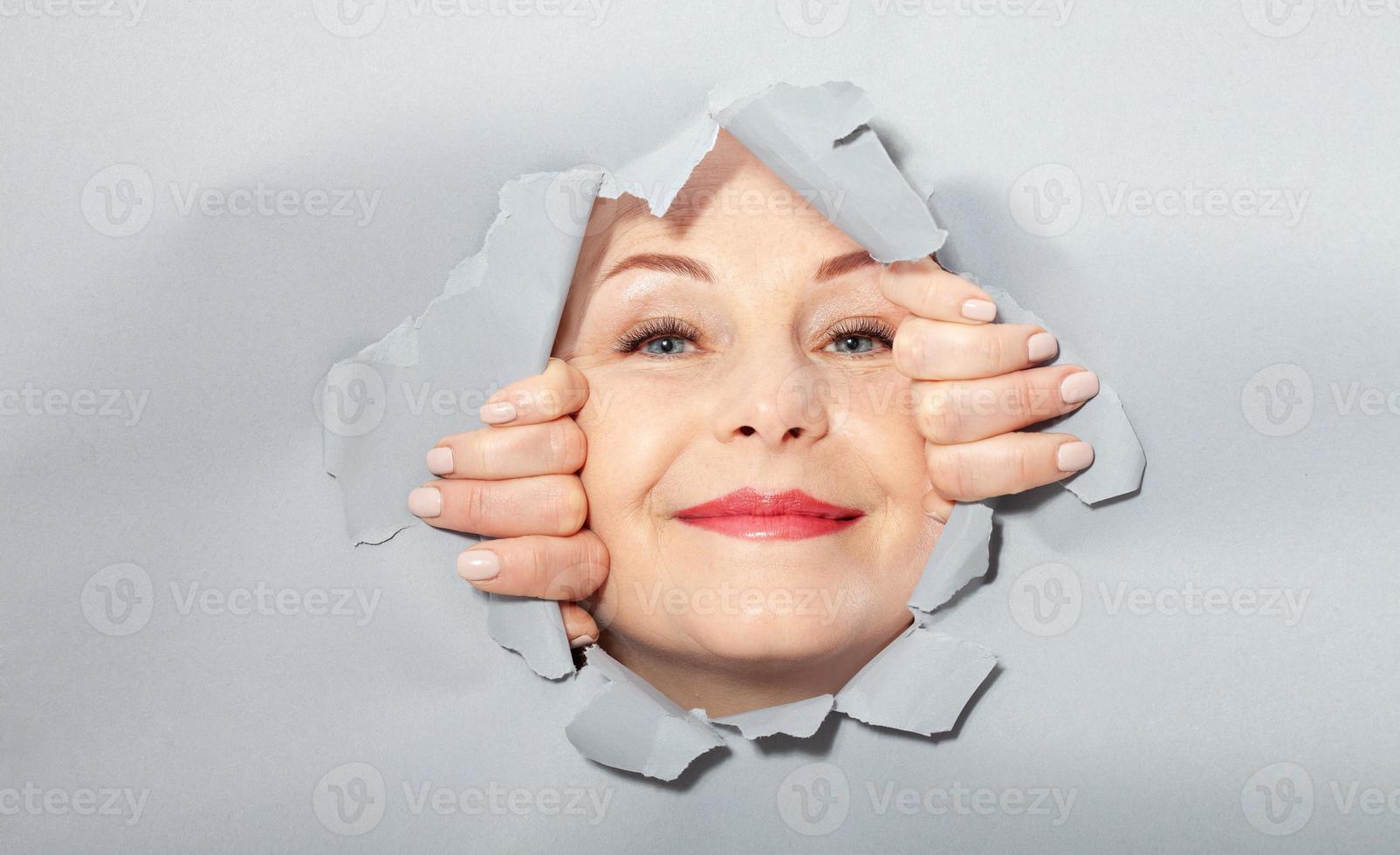 Surprised woman looking playfully in torn paper hole, has excited cheerful expression, looks through breakthrough of gray background. Wow, great news photo