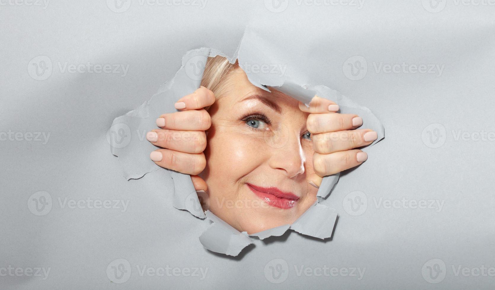 Surprised woman looking playfully in torn paper hole, has excited cheerful expression, looks through breakthrough of gray background. Wow, great news photo