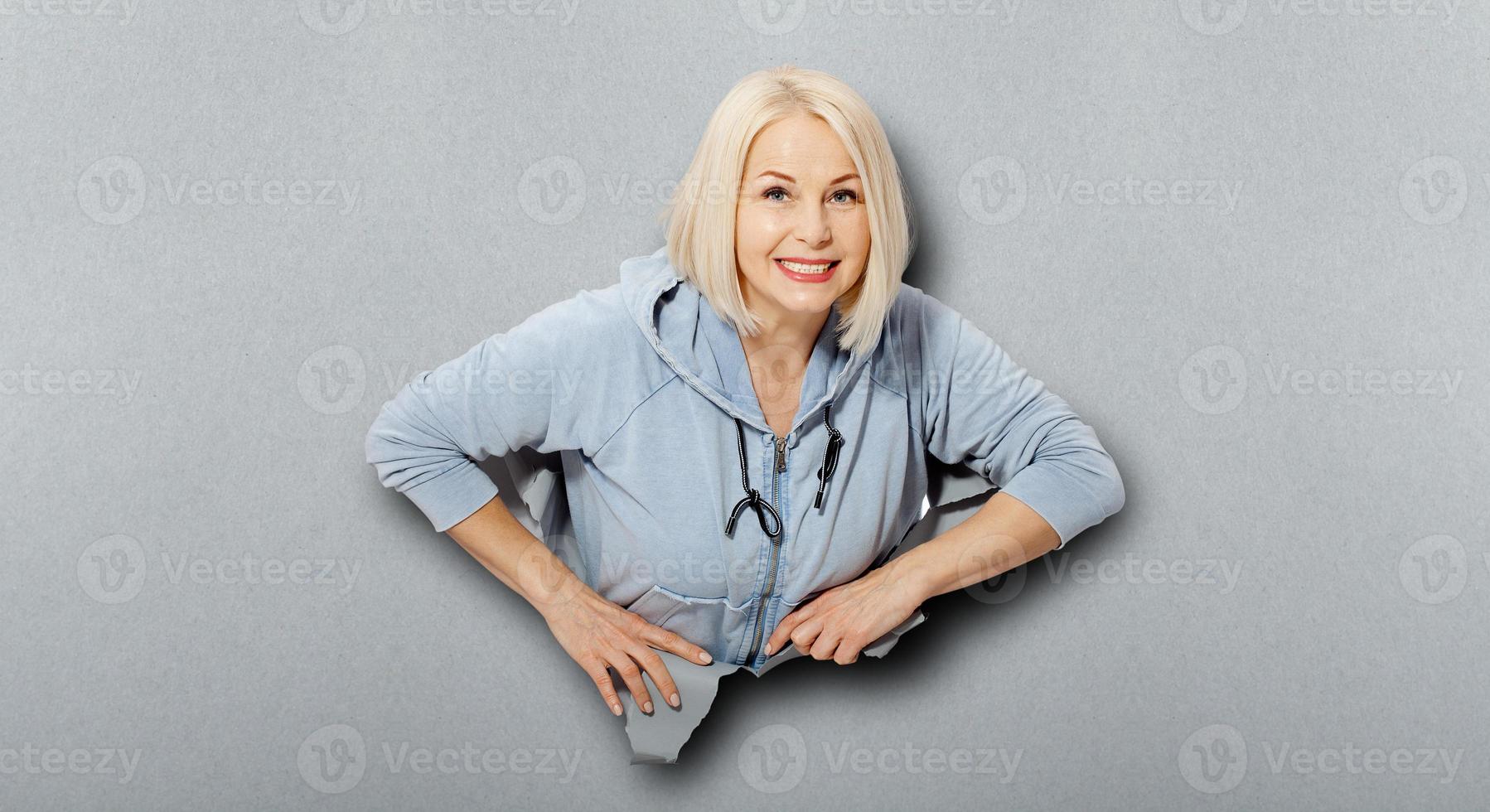 carismática mujer alegre con peinado rubio, con aspecto juguetón, viste con capucha de moda, se para en un agujero de papel roto, se da cuenta de que es divertido y agradable. fondo gris foto