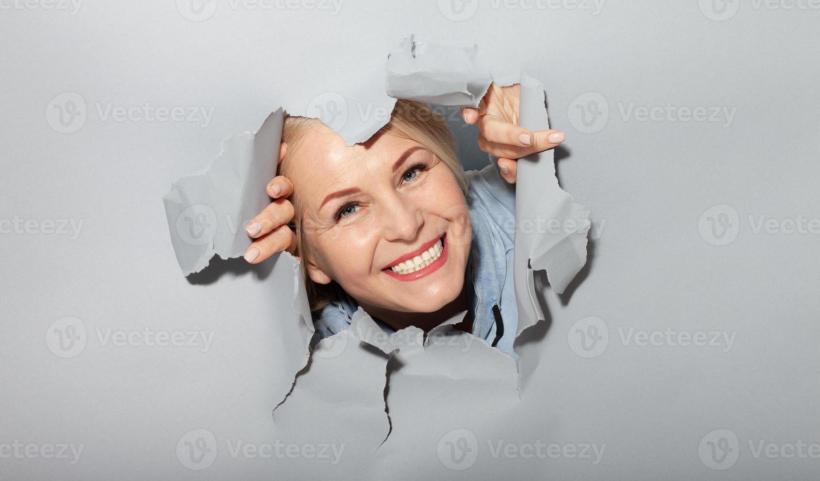 Surprised woman looking playfully in torn paper hole, has excited cheerful expression, looks through breakthrough of gray background. Wow, great news photo