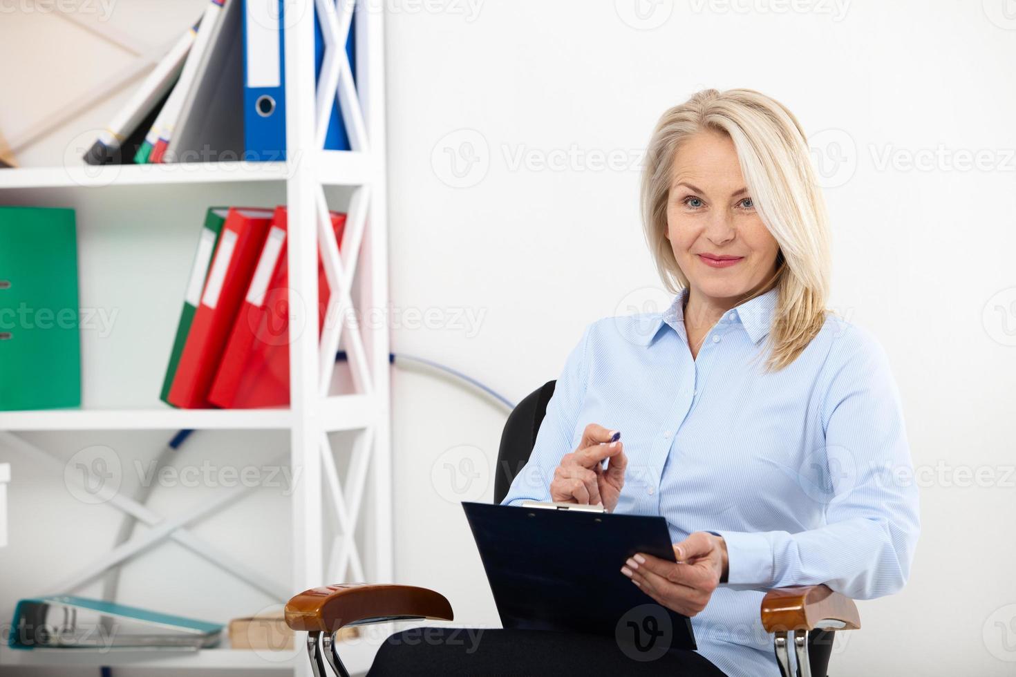 Business woman working in office with documents. Beautiful middle aged woman looking at camera with smile while siting in the office. photo