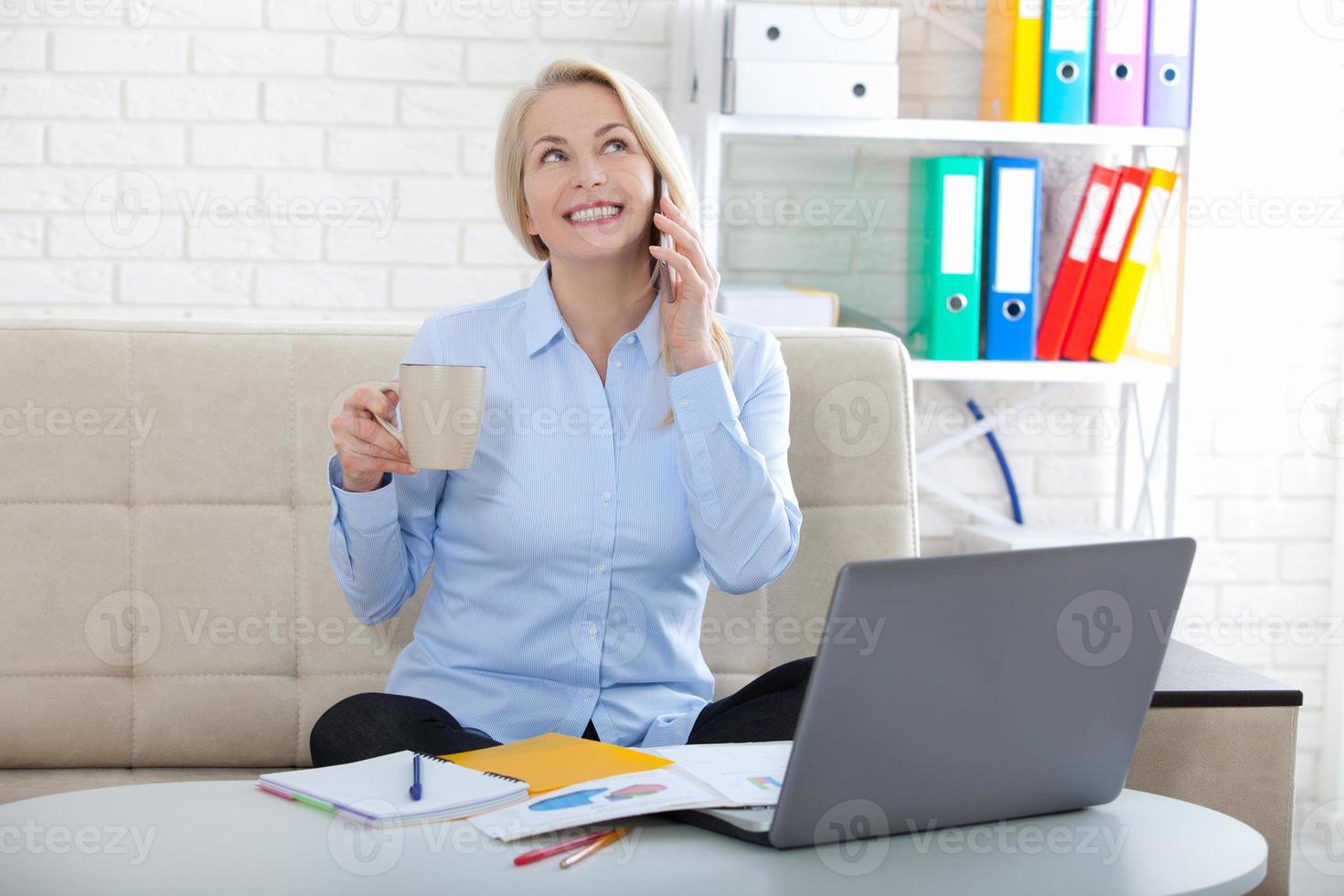 Listening to clients requirements. Beautiful middle aged woman with glasses talking on smart phone and smiling at her working place siting in the office. photo