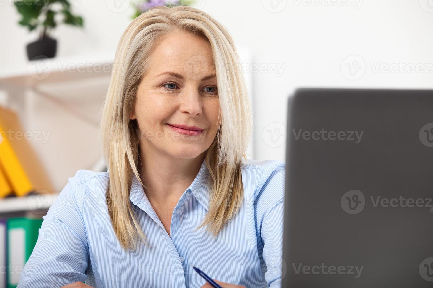 Working on new project. Business woman working in office with documents. Beautiful middle aged woman looking at laptop with smile while siting in the office. photo
