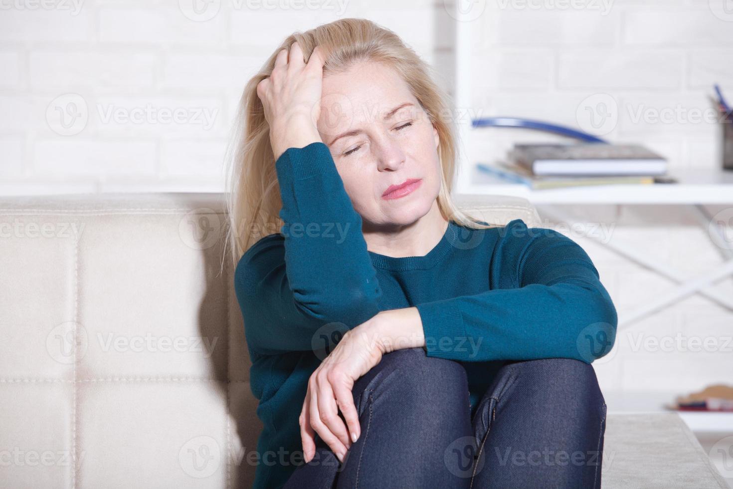 Sad depressed middle aged woman at home sitting on the couch, looking down and touching her forehead, loneliness and pain concept photo