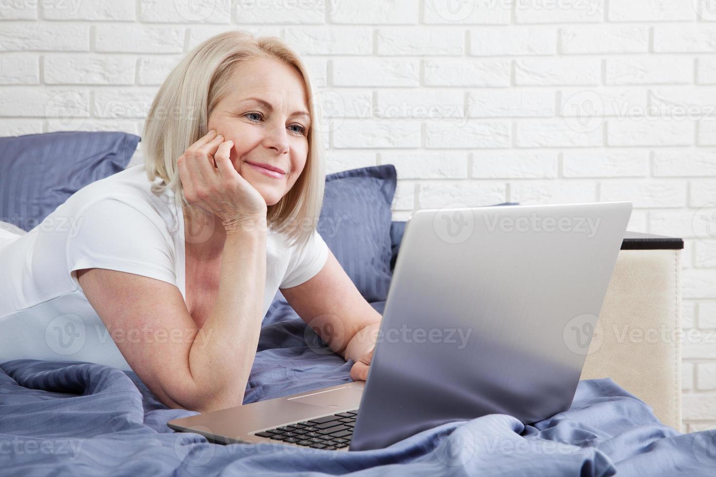 Smiling attractive woman lying on bed using laptop communicating online at home, happy girl typing on computer, enjoying internet shopping or chatting in social networks in bedroom. photo