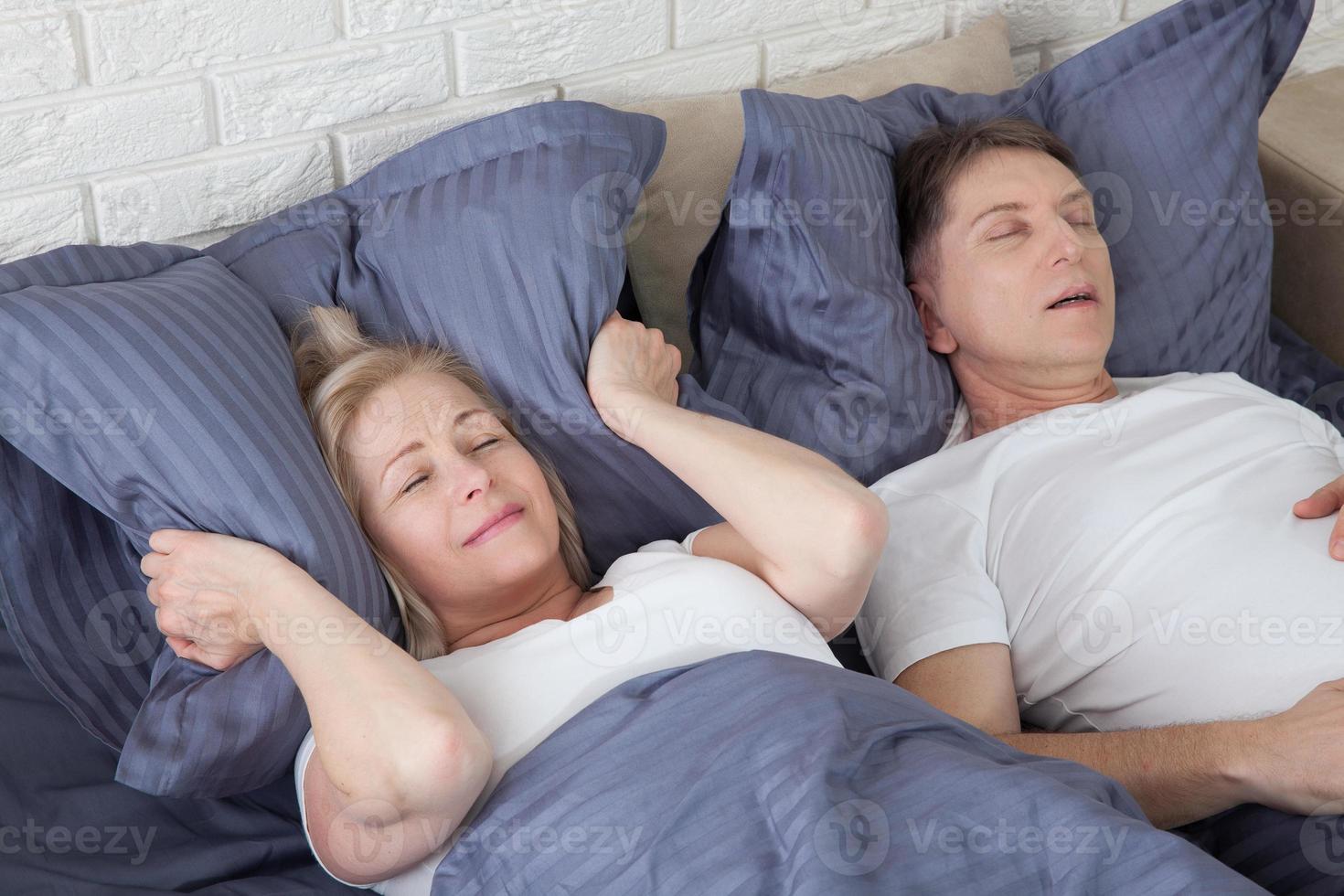Snoring man. Couple in bed, man snoring and woman can not sleep, covering ears with pillow for snore noise. Middle age couple in bed at home. photo